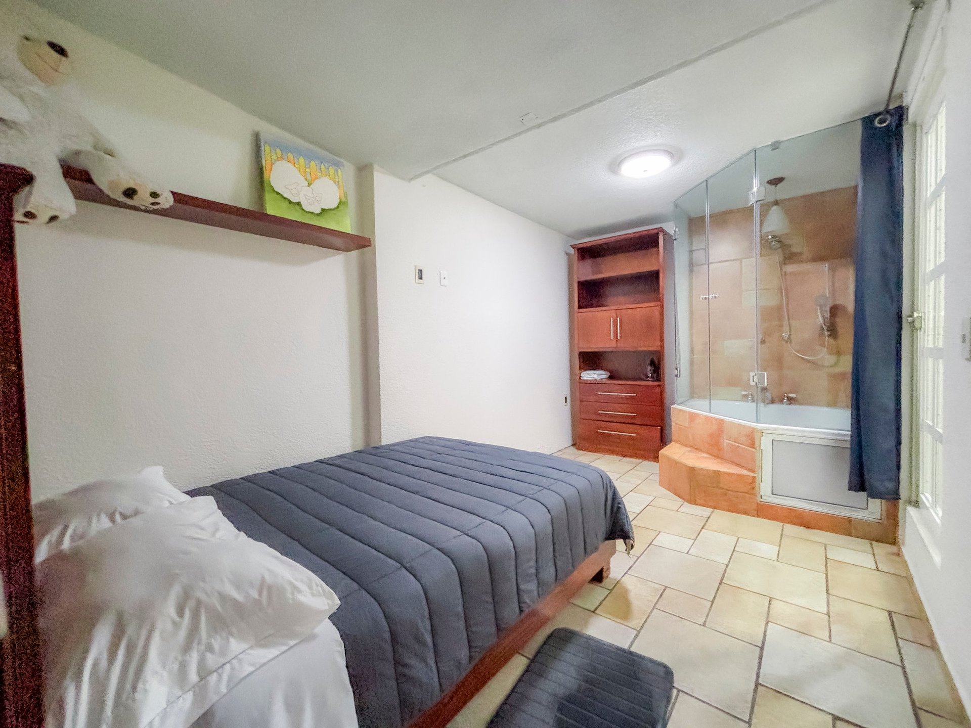 Modern bedroom with queen bed, ensuite bathroom with shower stall, wooden shelving, and tiled flooring.