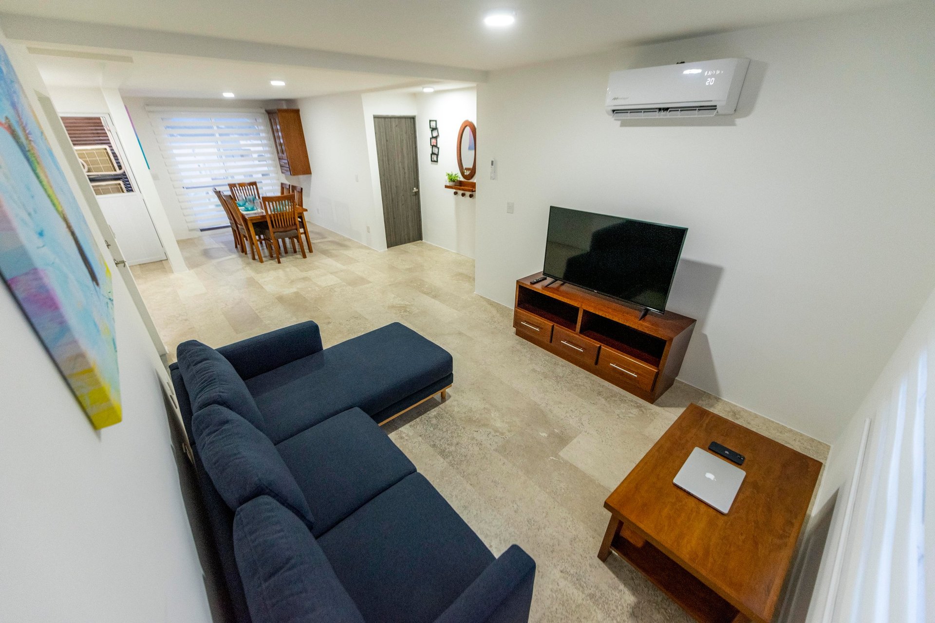 Modern living room with blue L-shaped sofa, wooden TV stand, and connected open-plan kitchen dining area with beige tile flooring