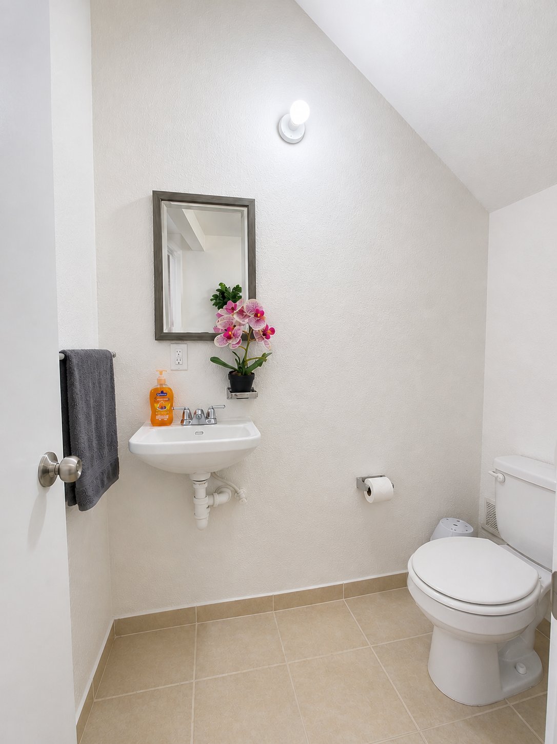 Modern bathroom with white walls, wall-mounted sink with mirror, pink flowers, gray towel, toilet, and beige tile flooring.