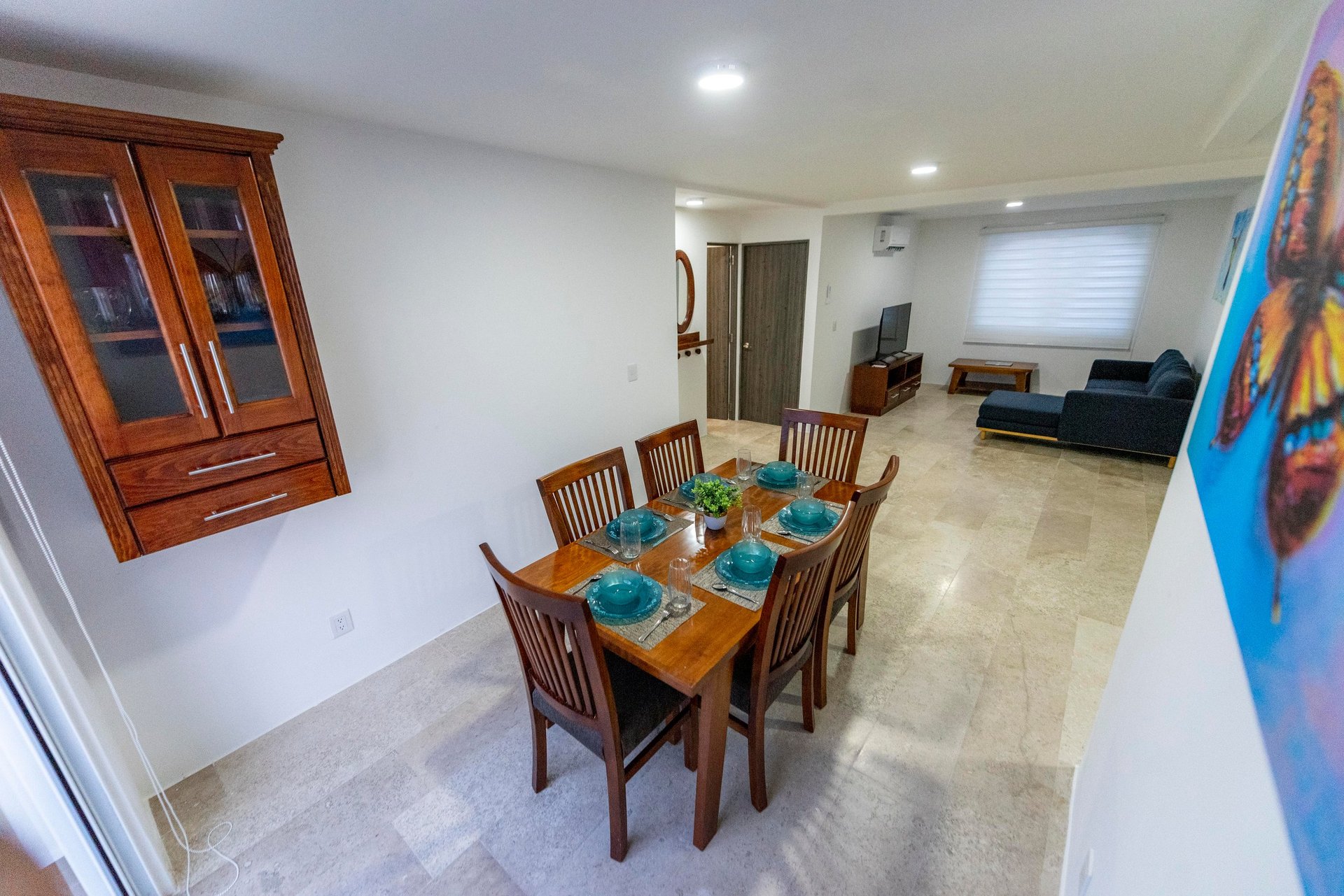 Open-plan dining and living room with wooden dining table set for four, kitchen cabinets visible, and living area with sofa in background