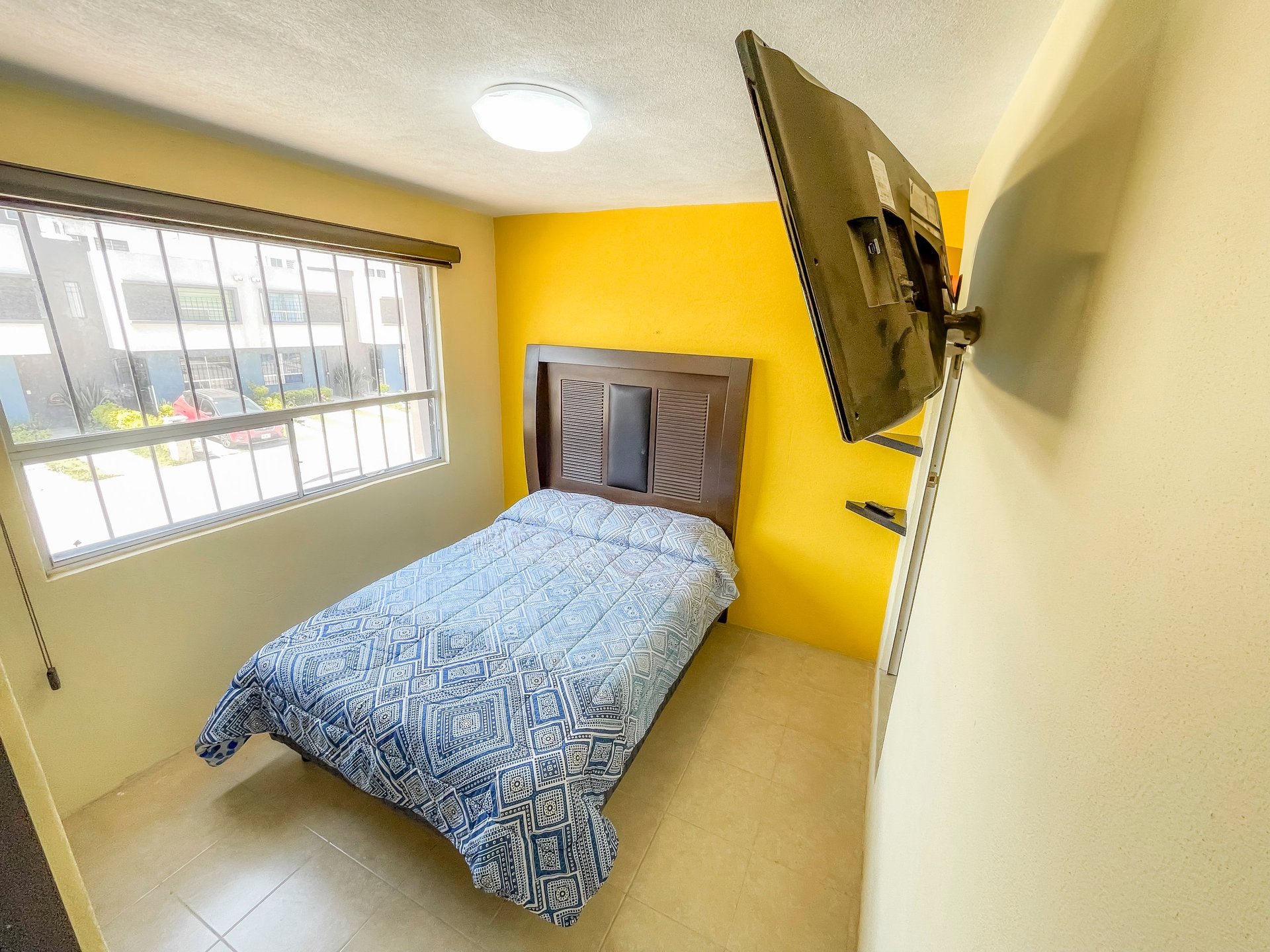 Small bedroom with yellow walls, twin bed with blue bedding, mounted TV, and barred window with natural light