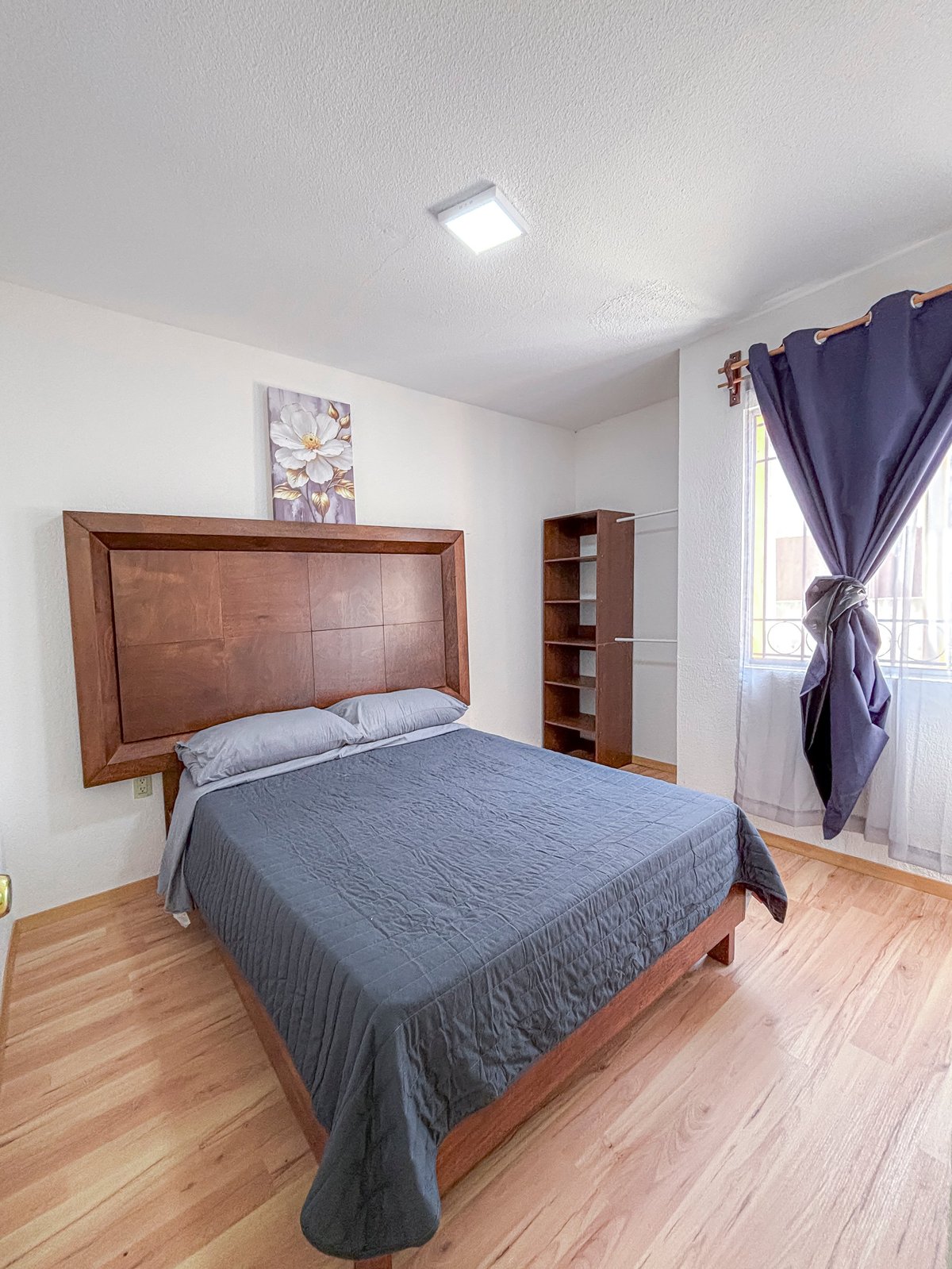 Modern bedroom with wooden bed frame, blue bedding, brown shelving unit, and purple curtains by window on hardwood floor