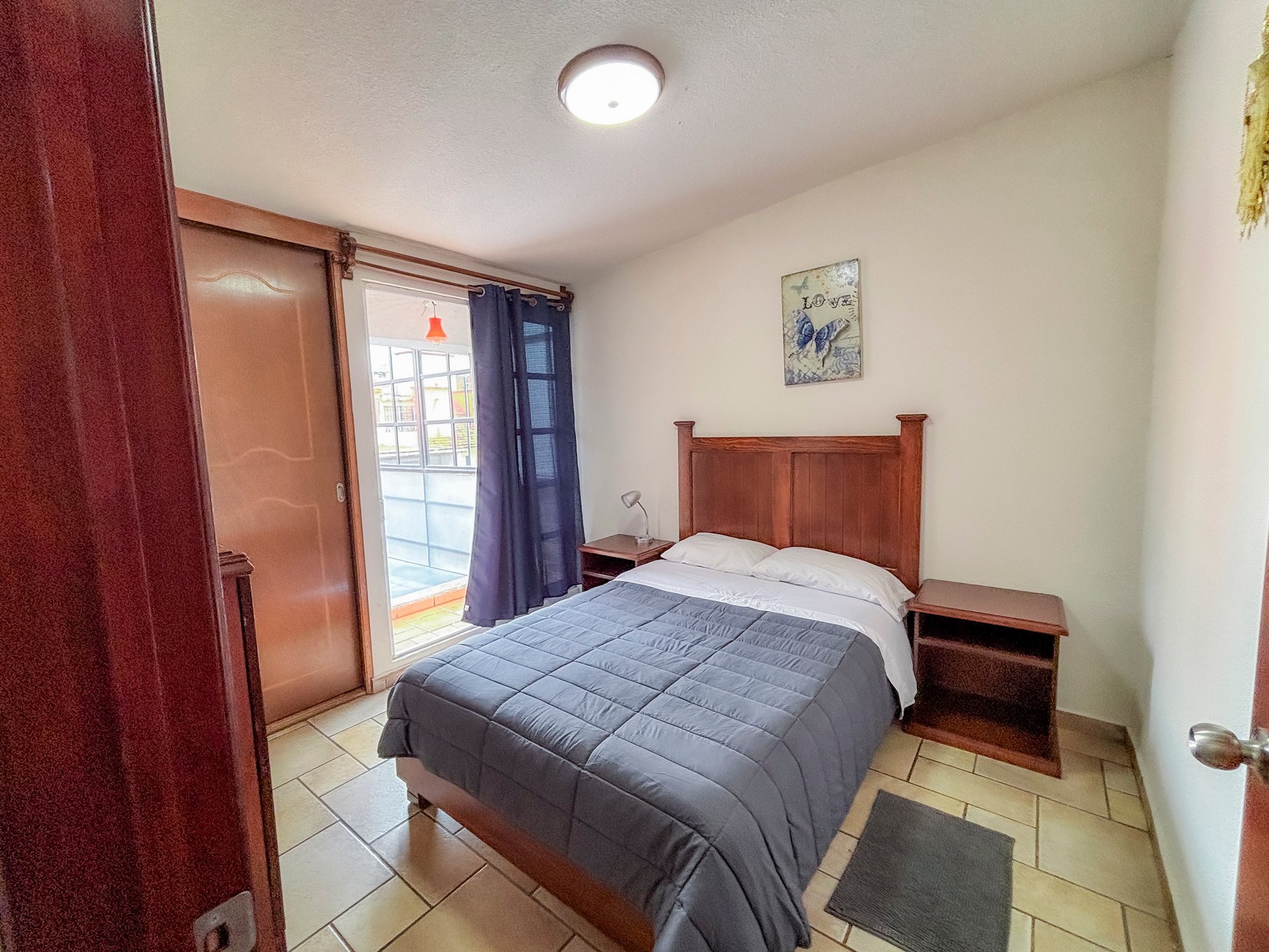 A small bedroom with a wooden bed, blue comforter, nightstands, and glass door to patio with blue curtains.