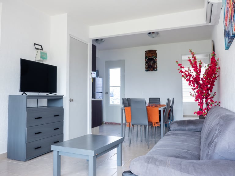Modern open-plan living room with gray furniture, TV on dresser, dining area with orange chairs, and red floral decoration