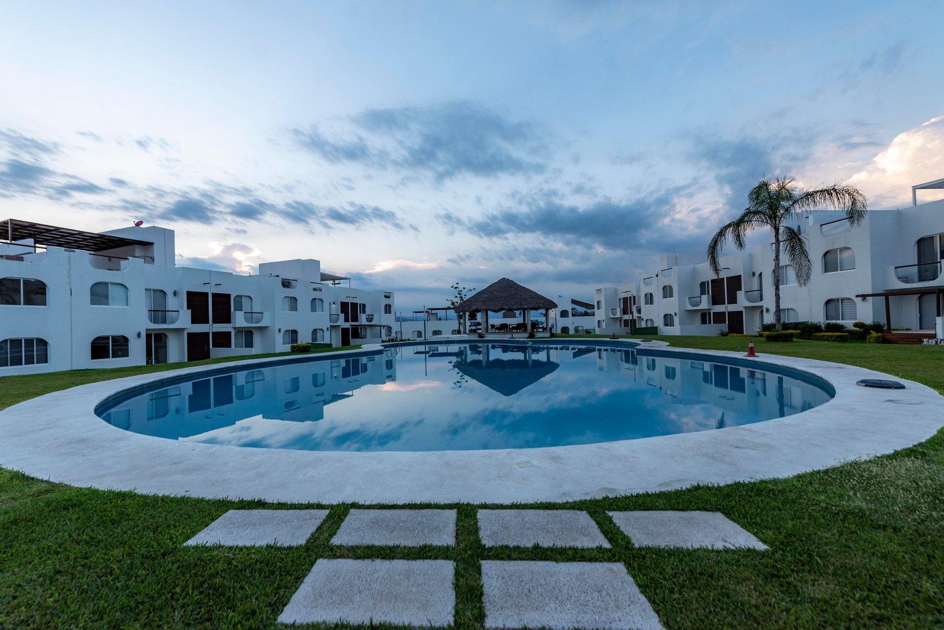 Oval swimming pool surrounded by white residential buildings at dusk with manicured grounds and palm trees
