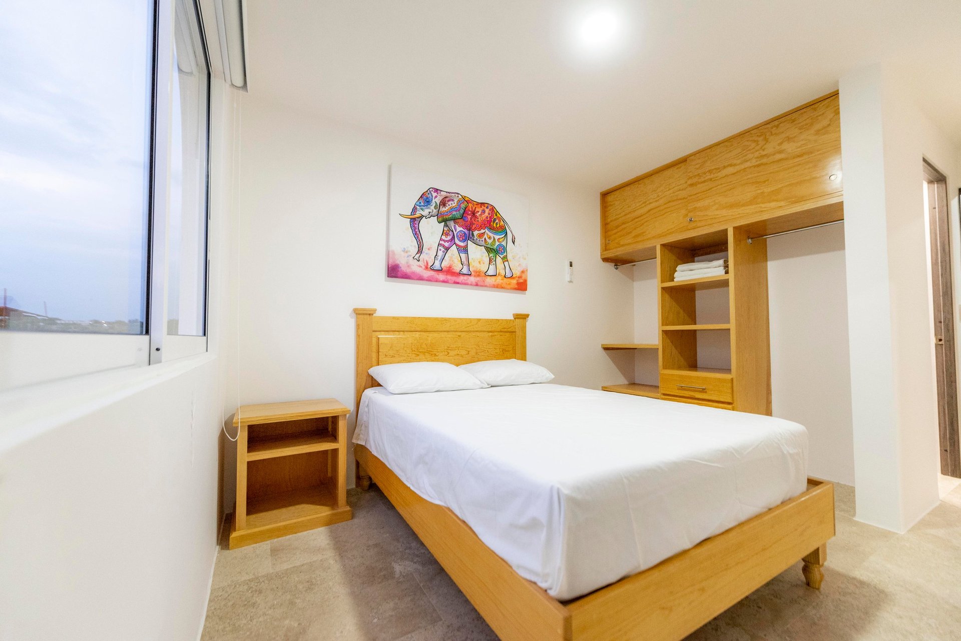 Modern bedroom with wooden bed, white linens, built-in wooden shelving, nightstand, and window with city views