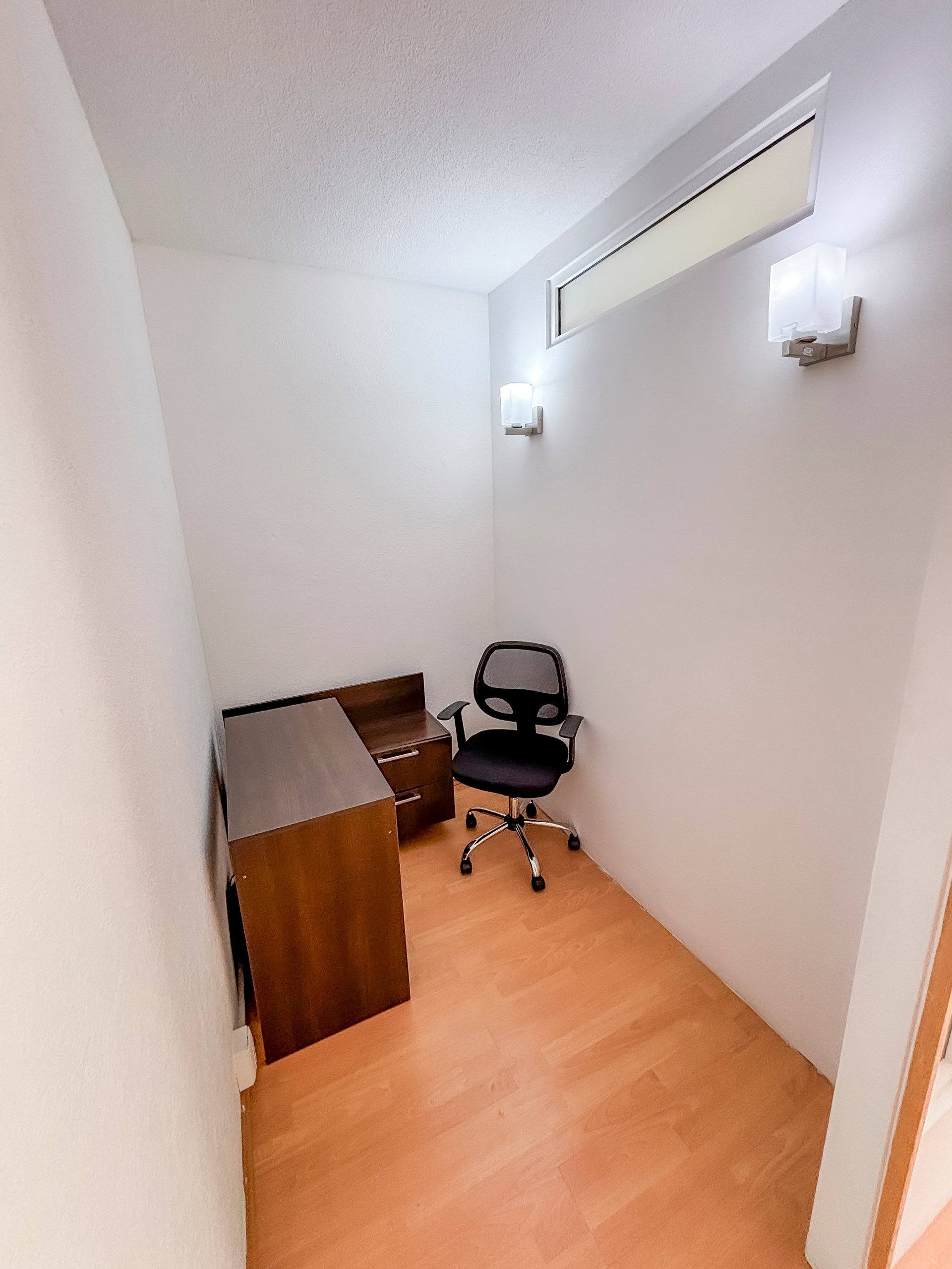 Small office nook with wooden desk, black mesh chair, and high window on white walls