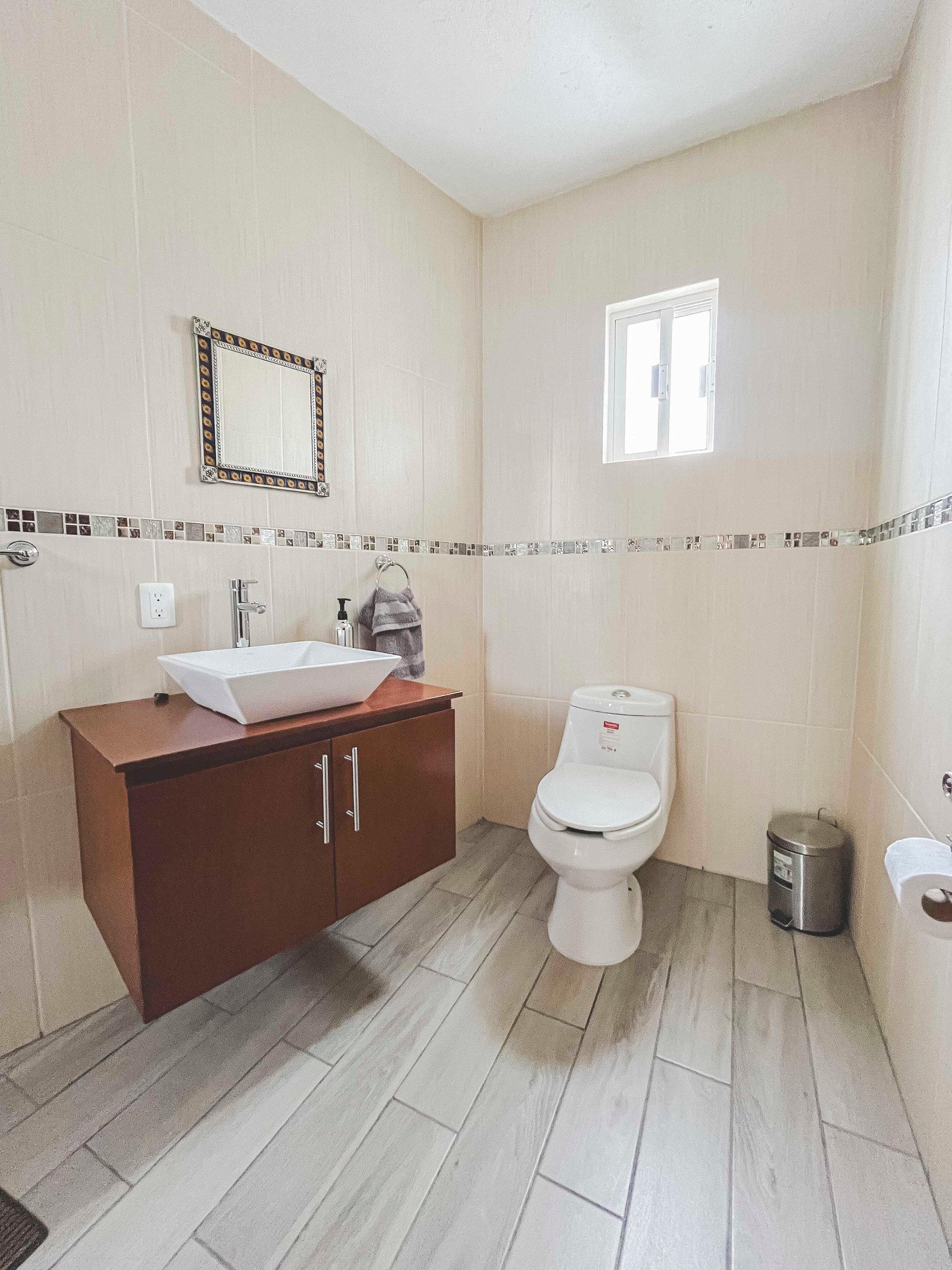 Modern bathroom with beige walls, white vessel sink on wood vanity, toilet, gray tile flooring, and small window