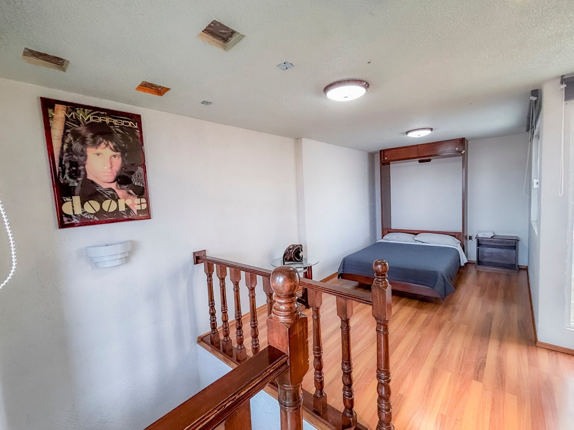 Spacious bedroom with wooden staircase, blue bed, The Doors poster, wooden flooring, and minimal furnishings.
