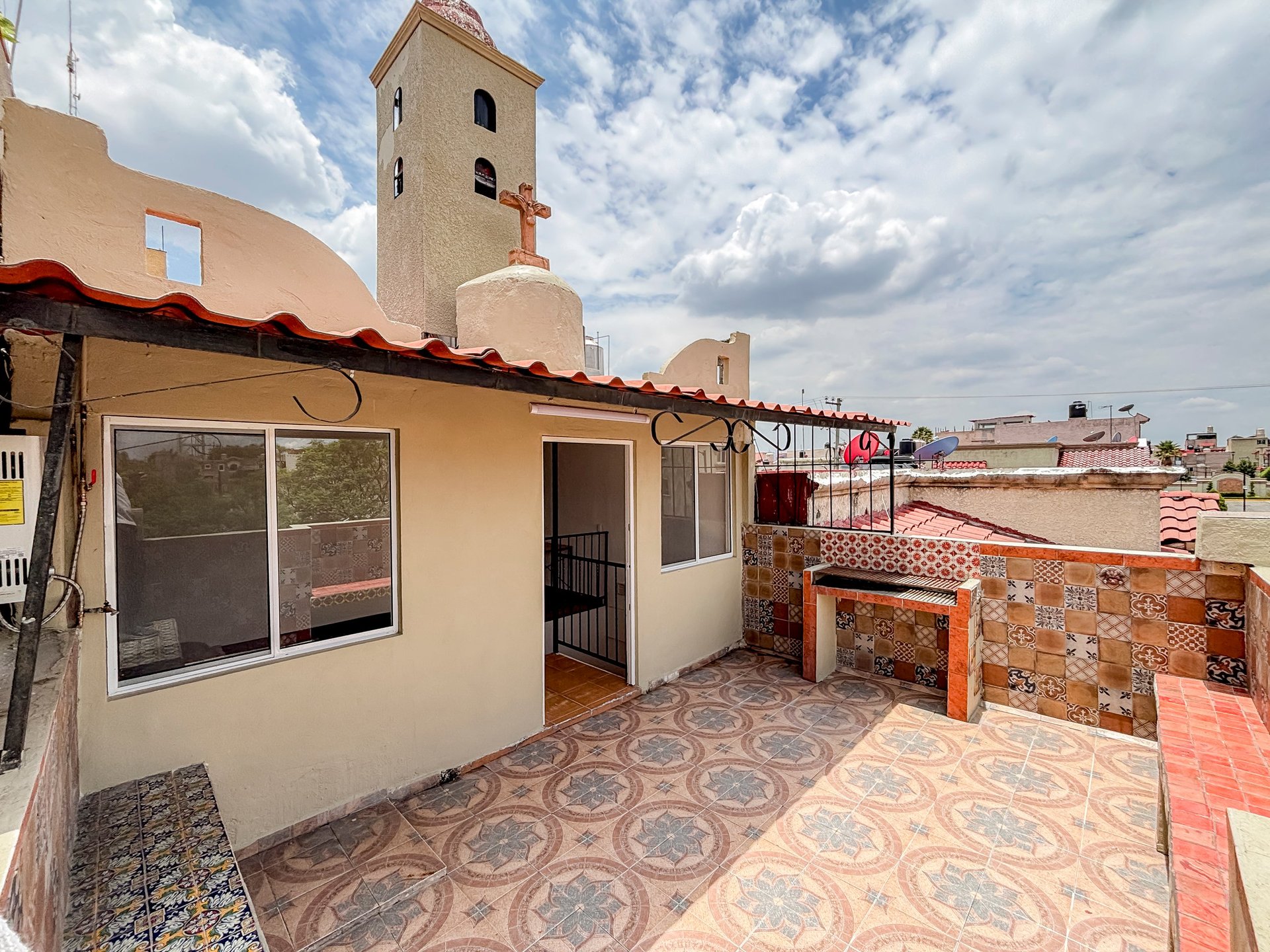 Terrace rooftop with cream-colored building, ornamental tile floor, red brick wall, and church tower visible in background under cloudy sky