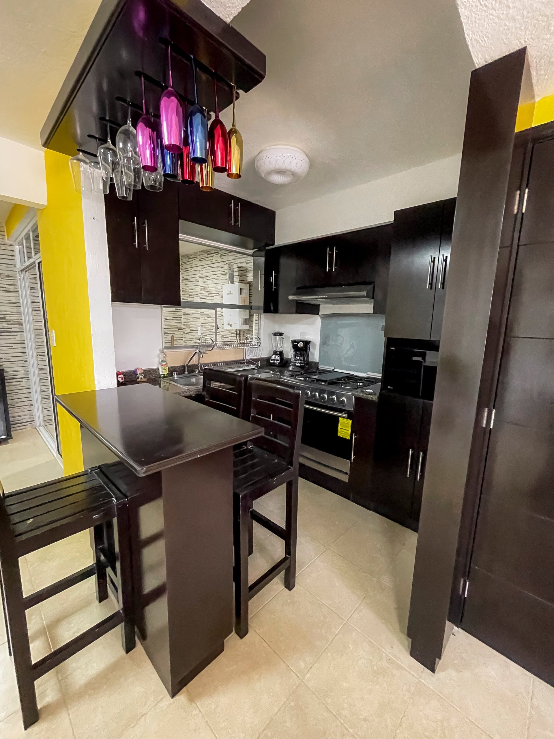 Modern kitchen with black cabinets, dark countertop island with bar seating, stainless steel appliances, and colorful hanging glasses above.