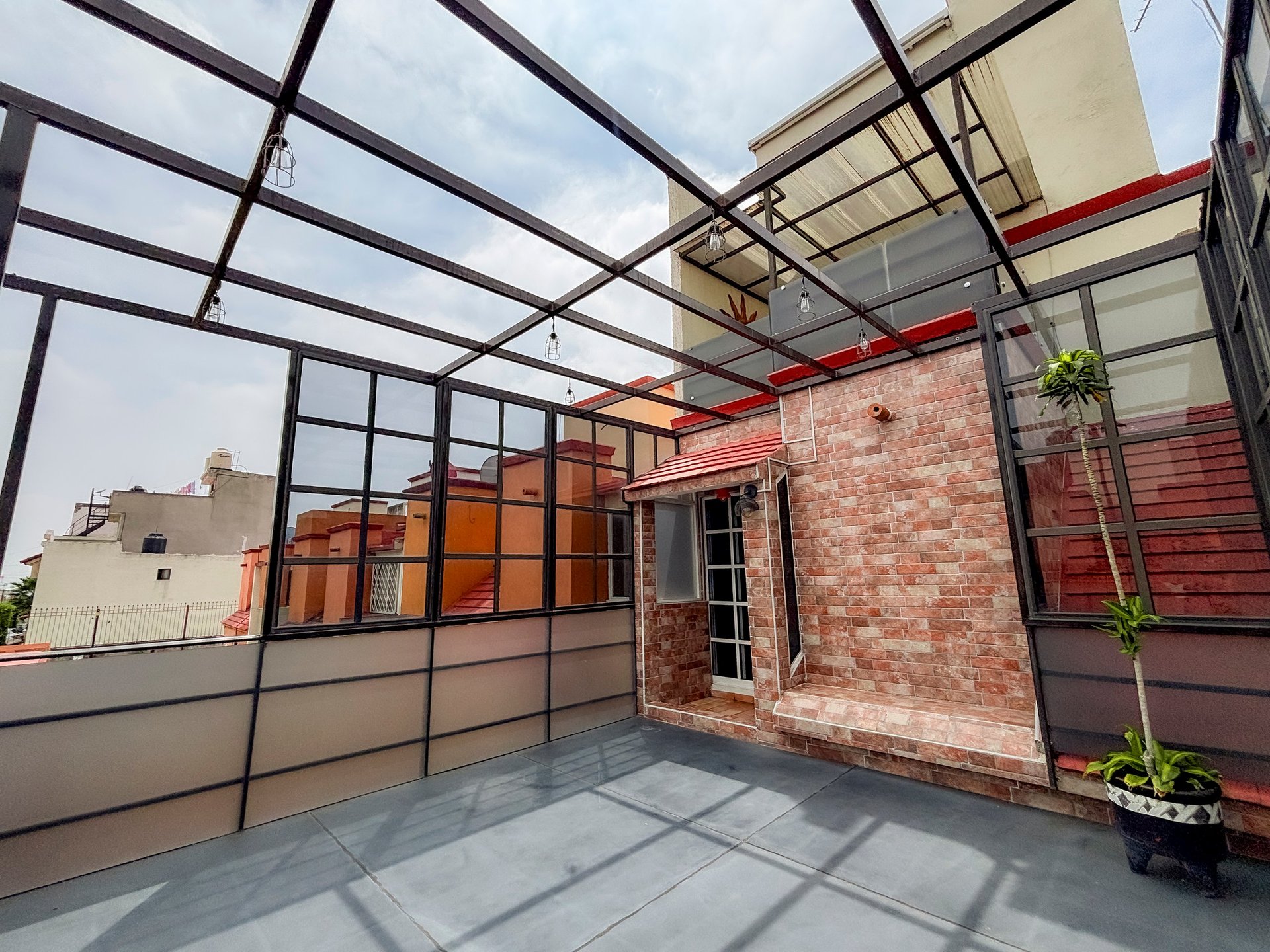 Modern rooftop terrace with glass and metal canopy, brick wall, and potted plants under natural lighting.