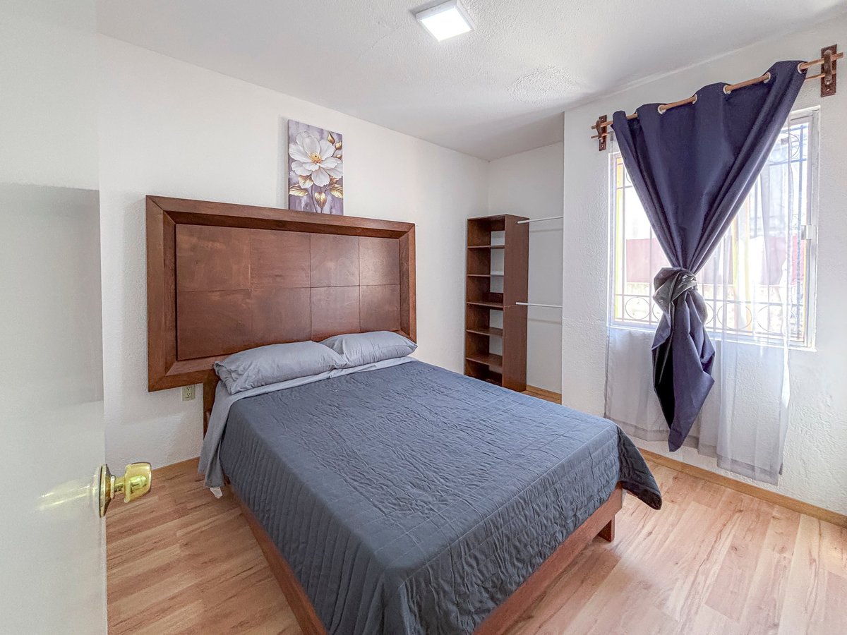 Bedroom with wooden bed frame, blue bedding, wooden nightstand, purple curtains, and light wood flooring.