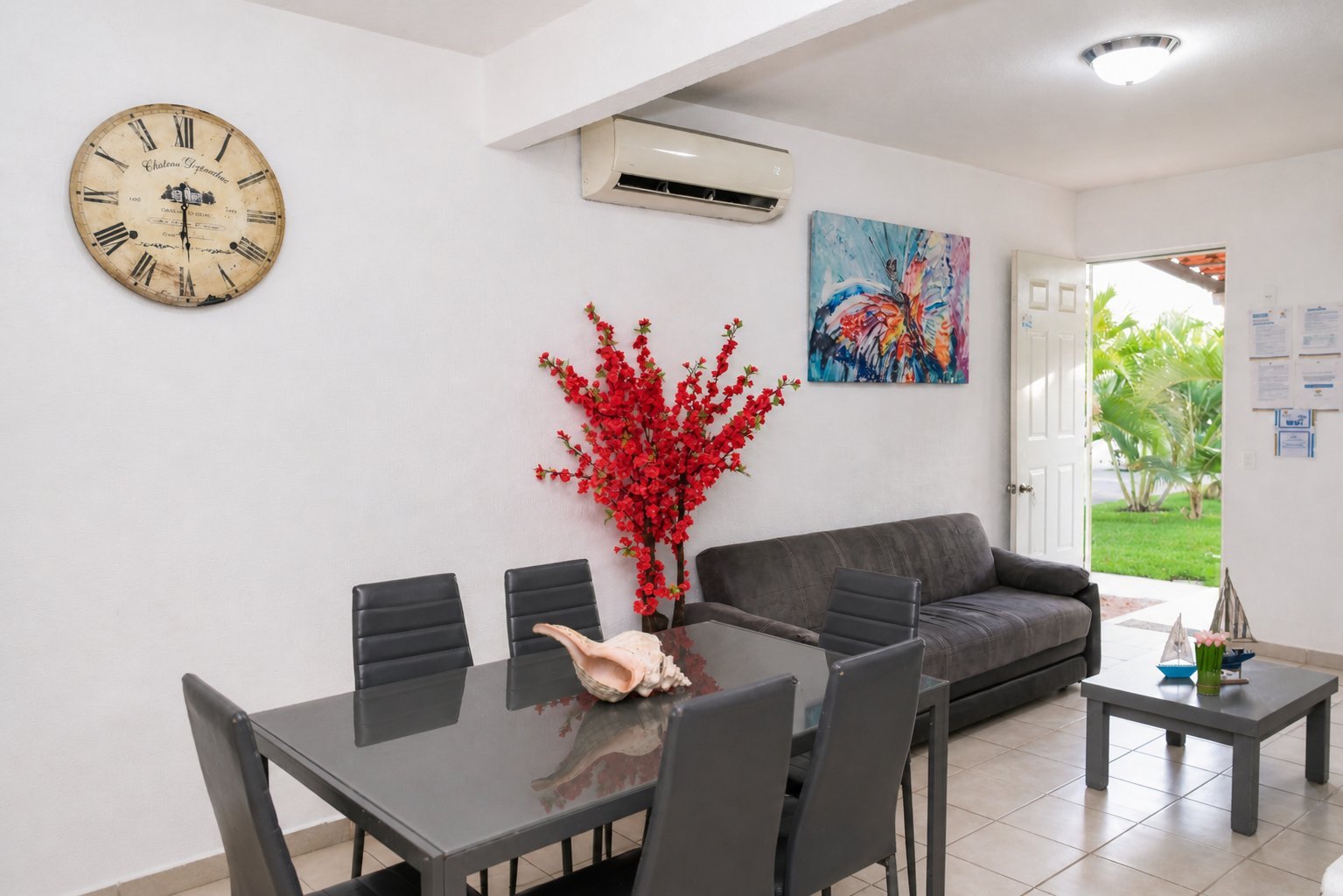 Modern living room with dining table, black sofa, red floral arrangement, wall clock, and view to garden.