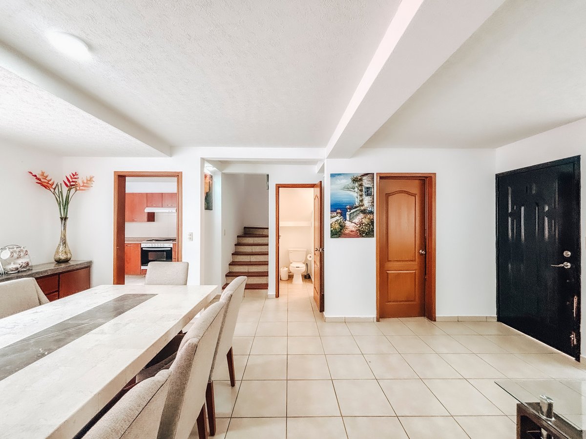 Modern kitchen with white counters, beige tile flooring, wooden doors, and open layout with staircase visible in background