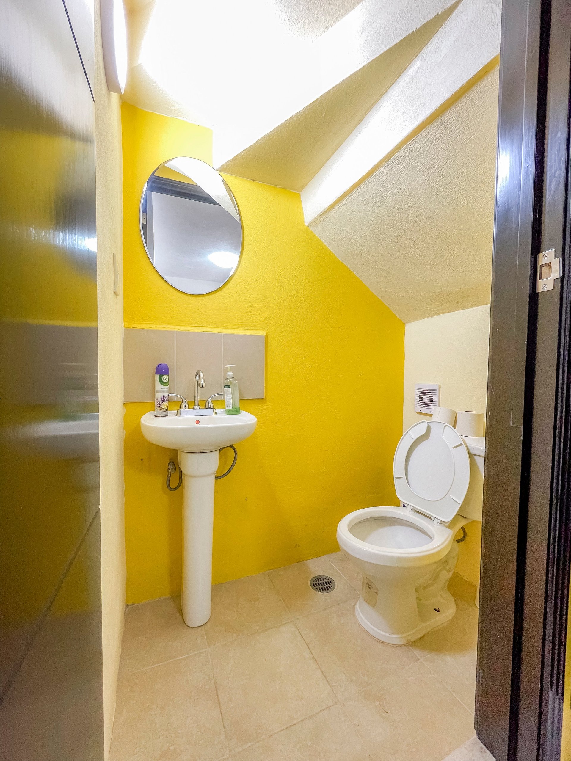 Modern bathroom with bright yellow walls, pedestal sink, round mirror, toilet, and sloped ceiling with skylight