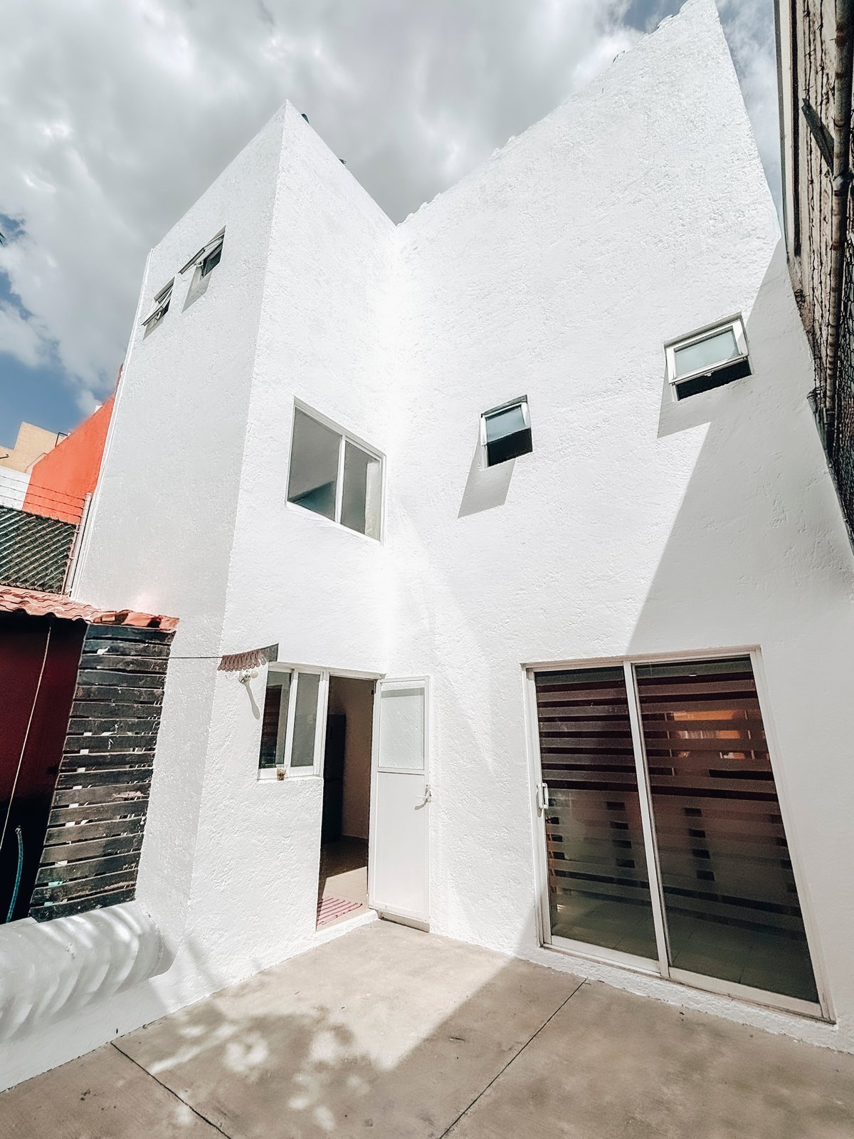 Modern white multi-story residential building with angular roofline, multiple windows, and brown wooden doors viewed from front courtyard