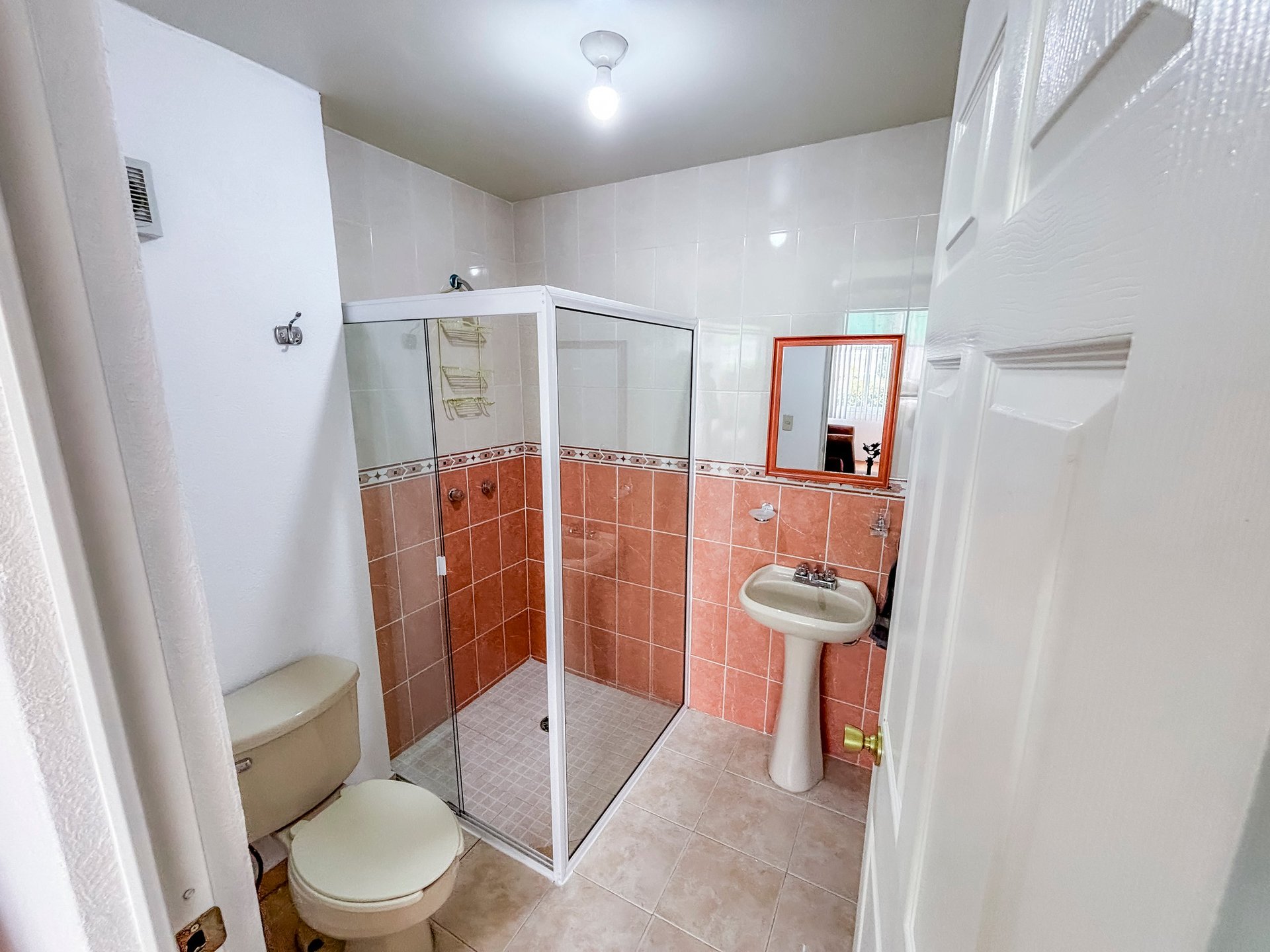 Modern bathroom with white walls, red tile accents, glass shower enclosure, toilet, pedestal sink, and orange-framed mirror