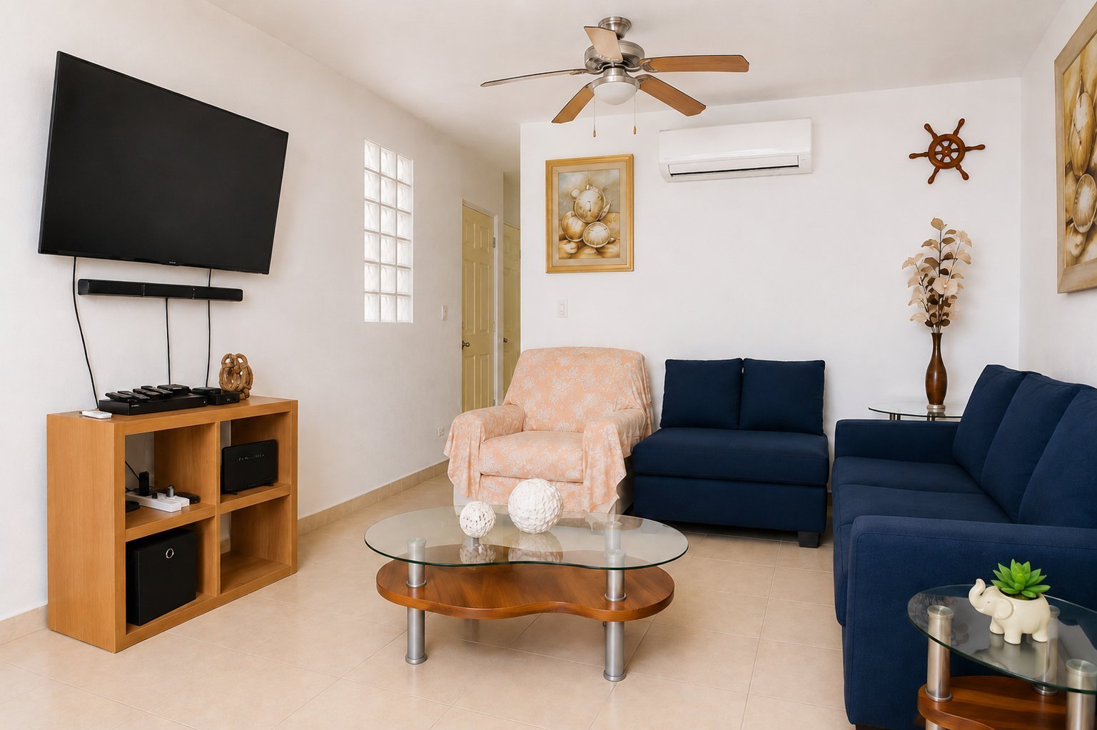 Modern living room with flat-screen TV on wooden stand, beige armchair, dark blue sofa, glass coffee table, and nautical decor