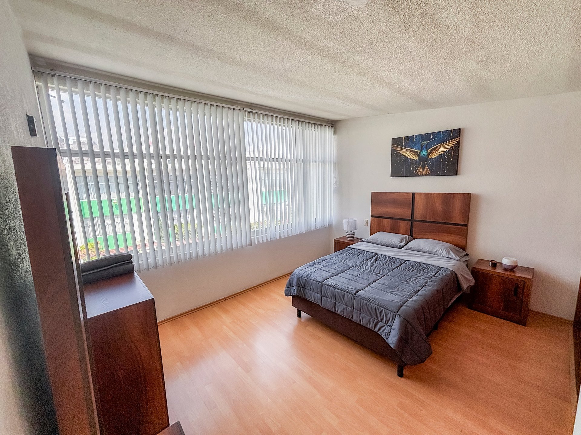 Spacious bedroom with wooden bed, nightstands, large window with vertical blinds, and hardwood flooring
