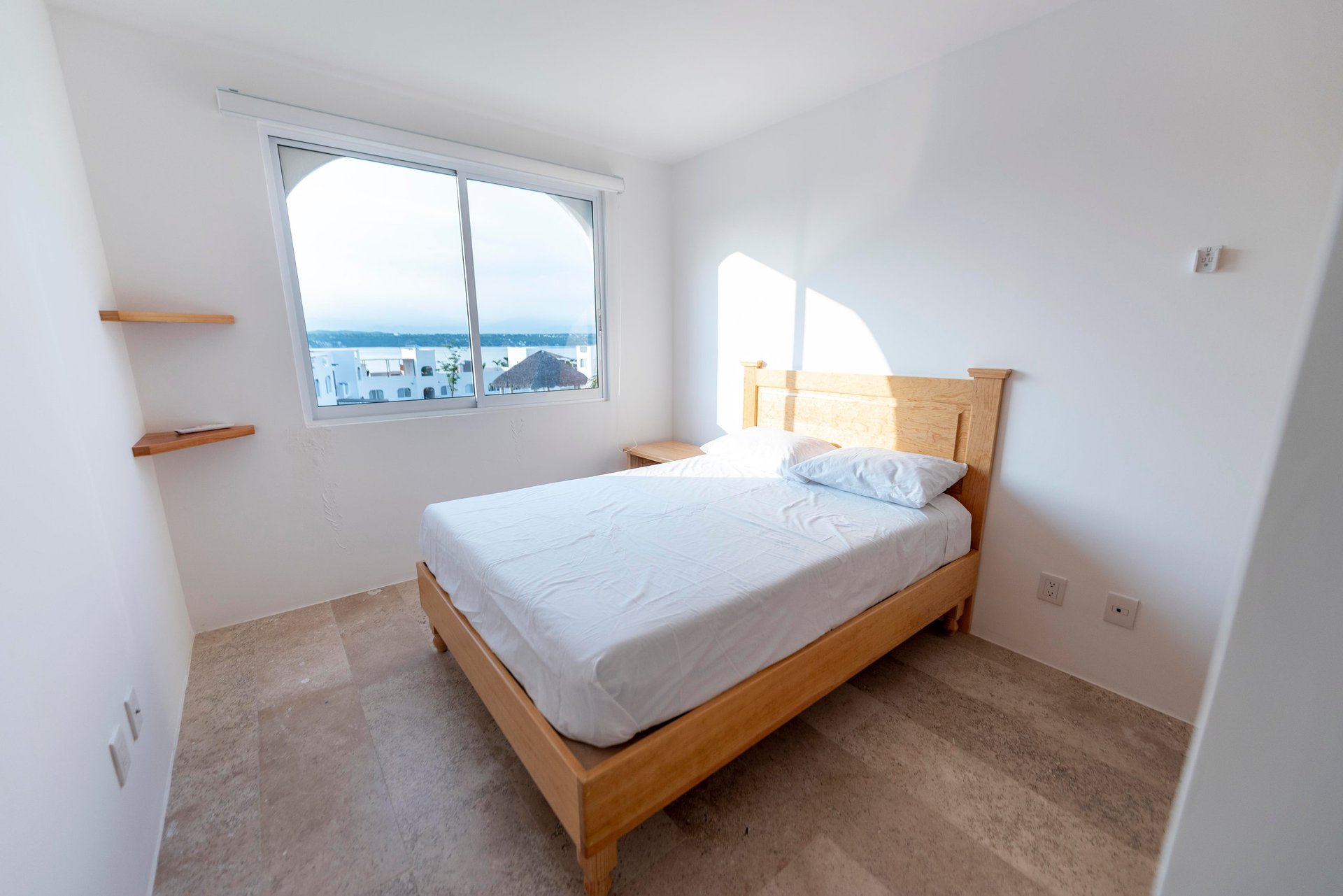 Bright modern bedroom with wooden bed, white linens, and large window overlooking city waterfront