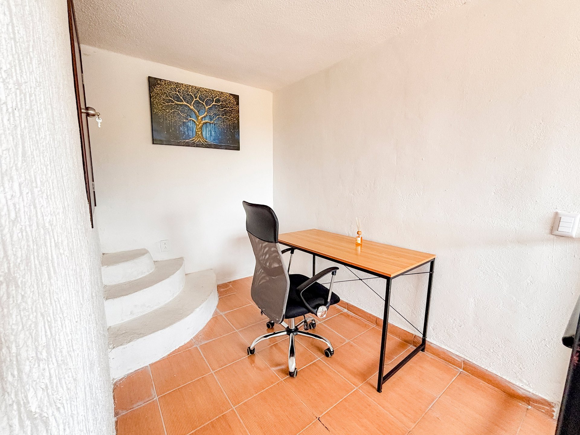 Small minimalist home office with wooden desk, black office chair, and framed artwork on white walls with terracotta tile flooring