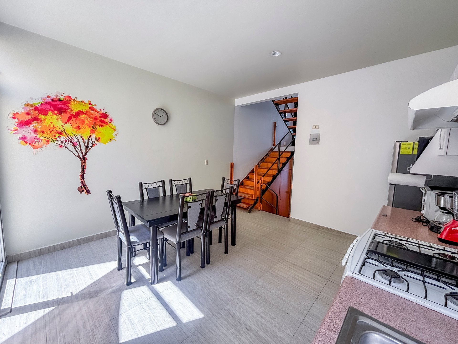Modern kitchen and dining area with black table and chairs, colorful flower wall art, and wooden staircase visible in background