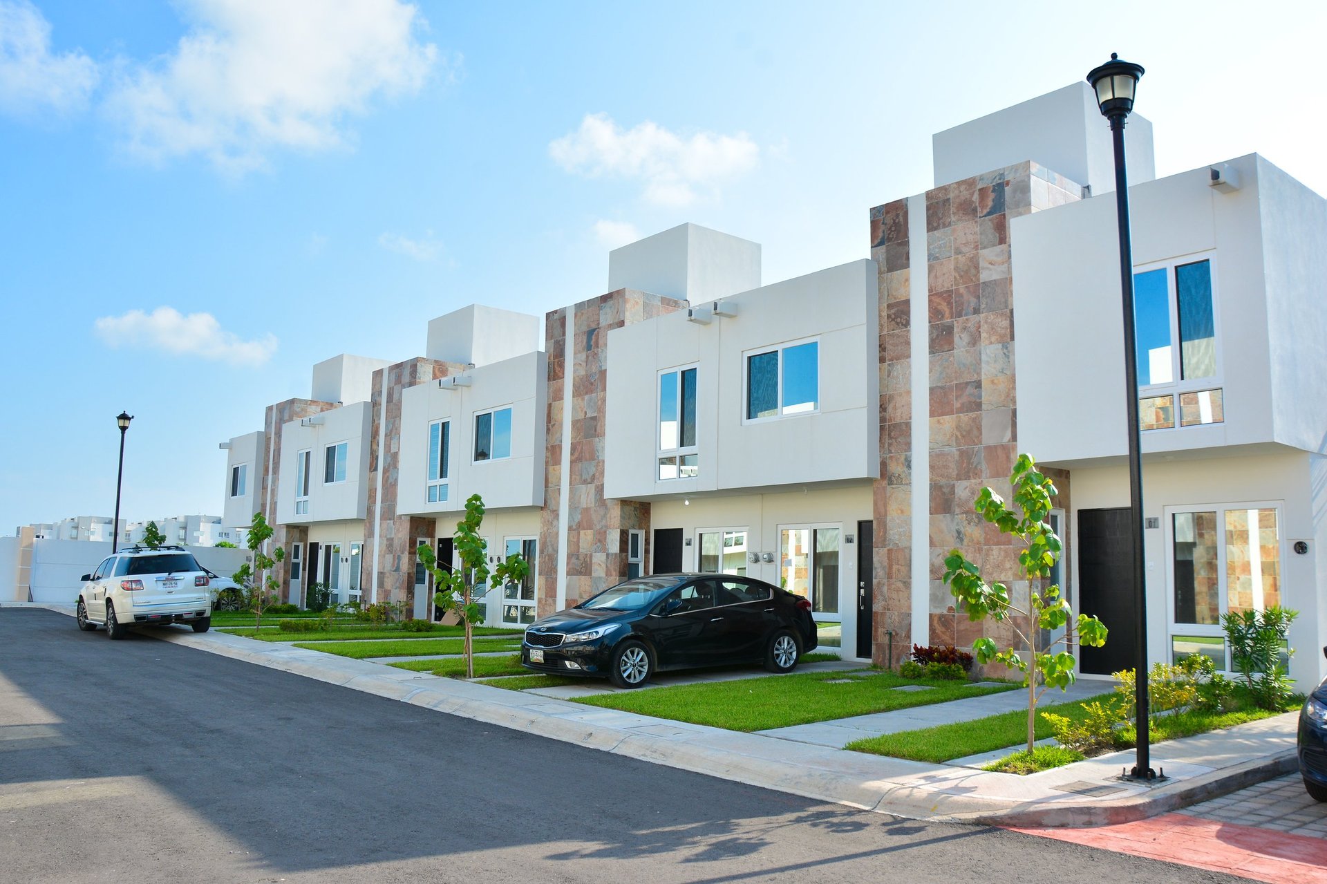Row of modern white townhouses with stone accents, parked cars in front, green lawns, and street lamps on a sunny day