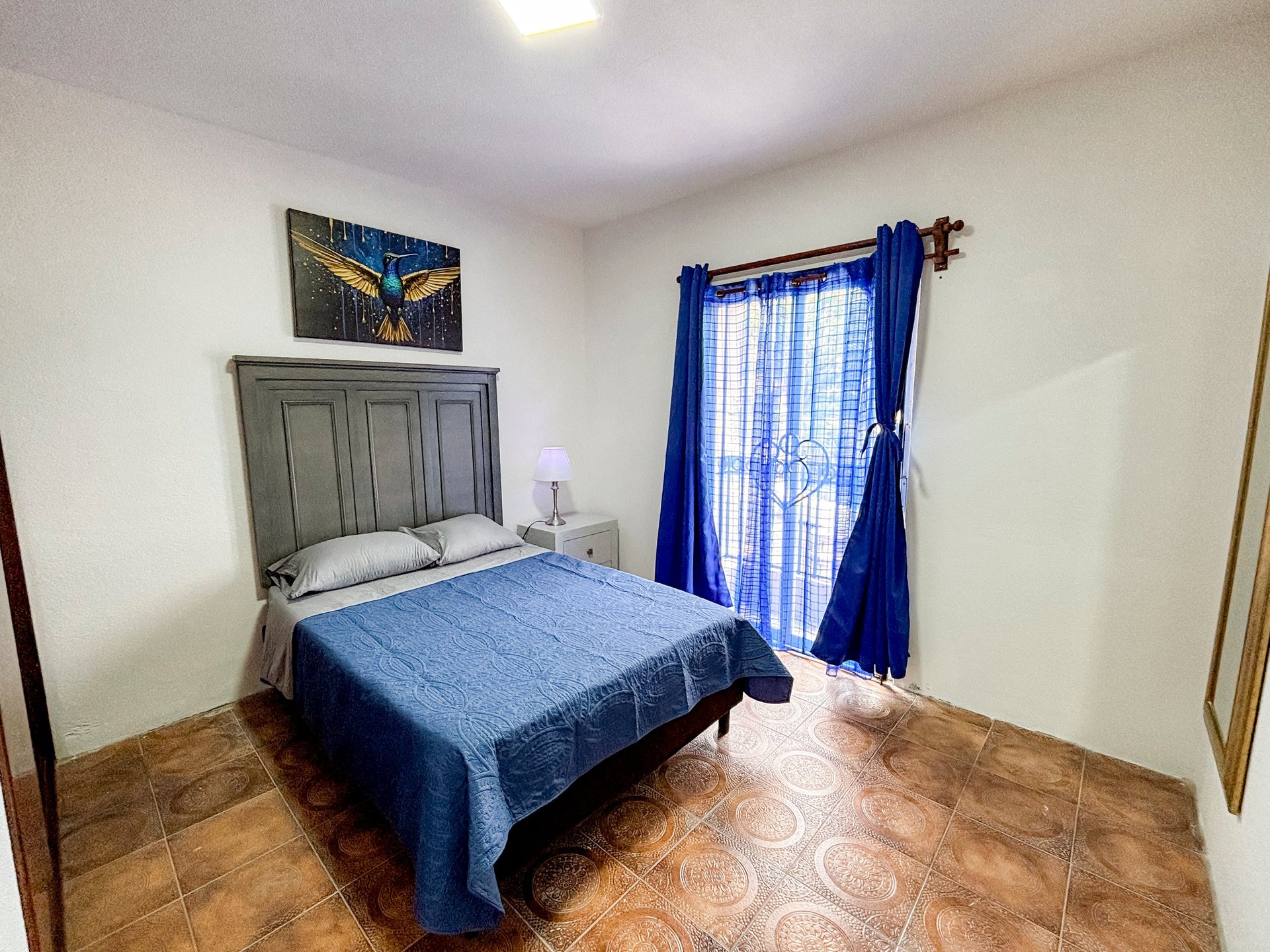 A bedroom with a blue bed, wooden headboard, nightstand, blue curtains on the window, and terracotta tile flooring.