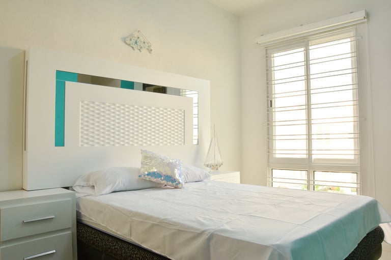 Modern bedroom with teal and white geometric headboard, white bedding, nightstand, and bright window with shutters