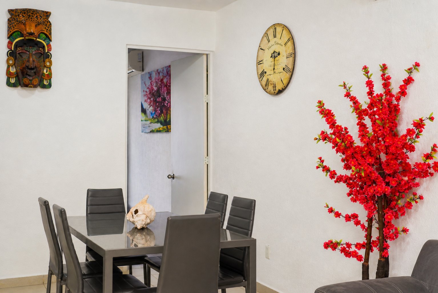 Modern dining room with black table and chairs, decorative red artificial tree, wall clock, religious icon, and fresh floral arrangement.