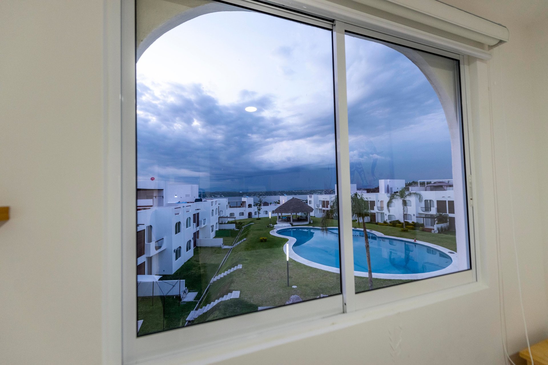 Large window view of residential buildings, swimming pool, and lawn with stormy sky and distant city skyline