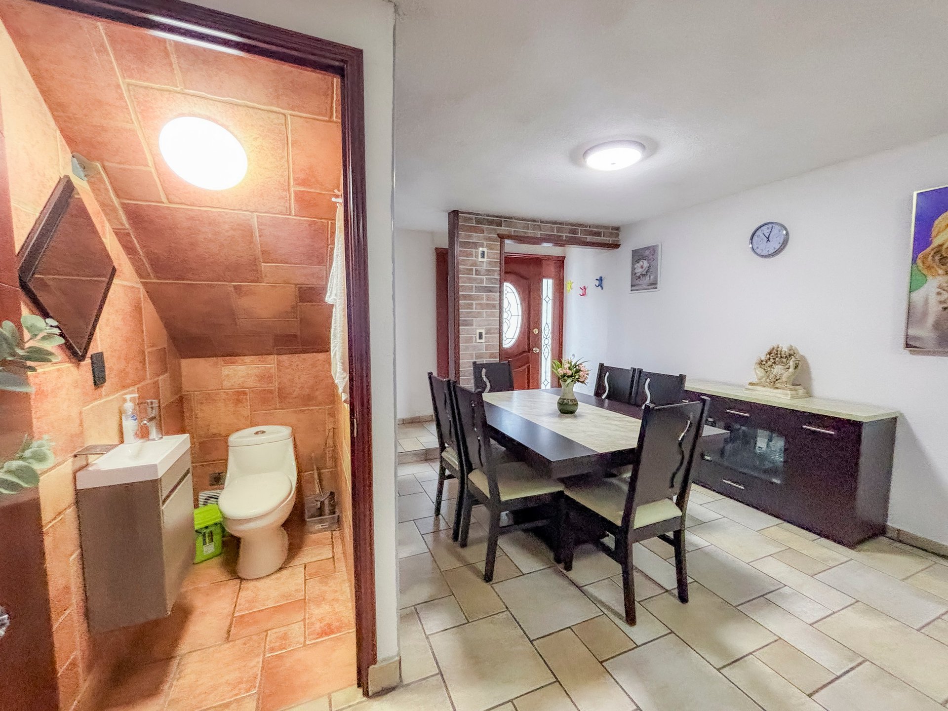 Open floor plan showing a bright dining area with black dining table and chairs, adjoining a bathroom with terracotta tile accents.
