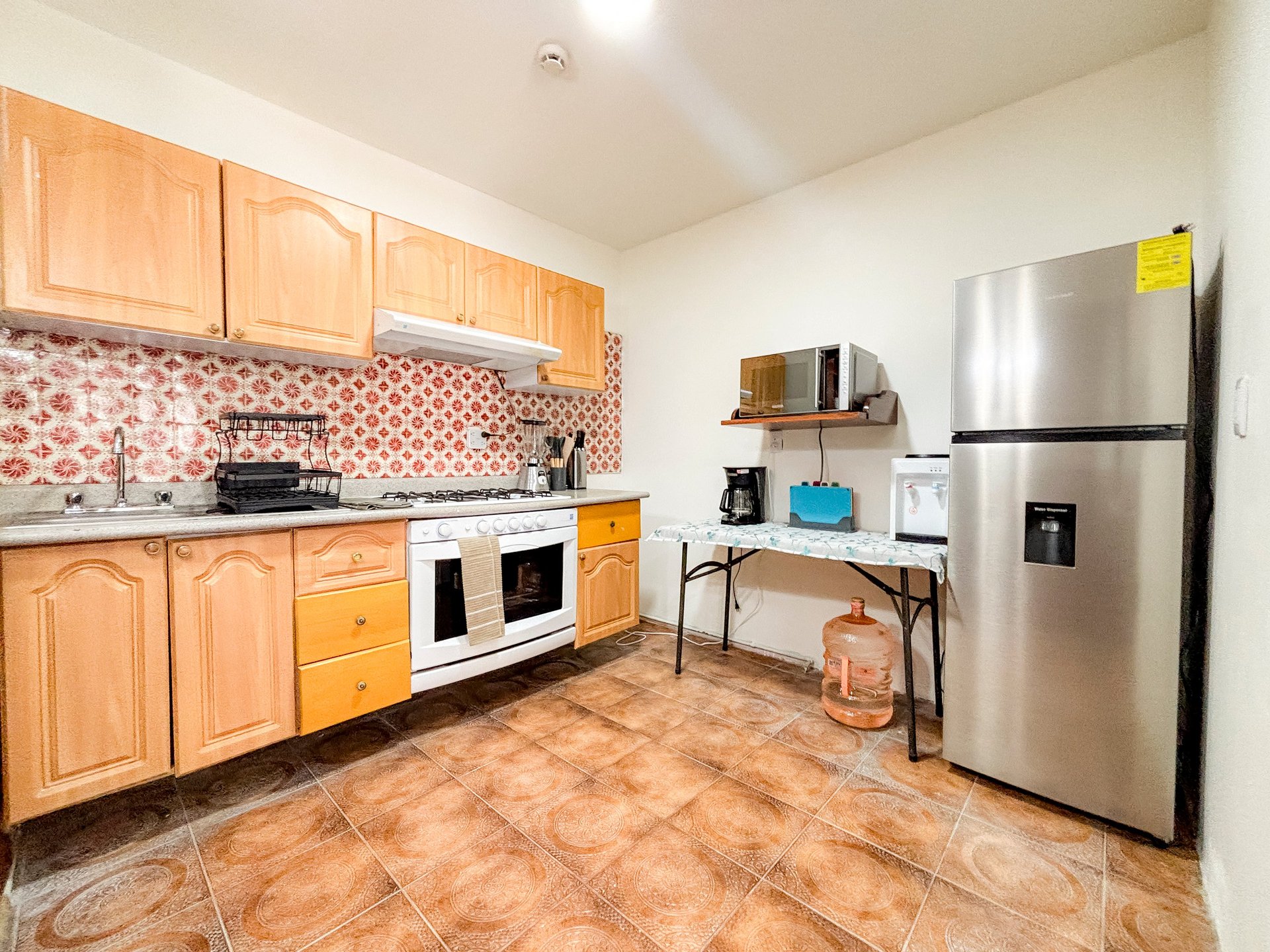 Spacious kitchen with wooden cabinets, red patterned backsplash, white stove, stainless steel refrigerator, and tile flooring
