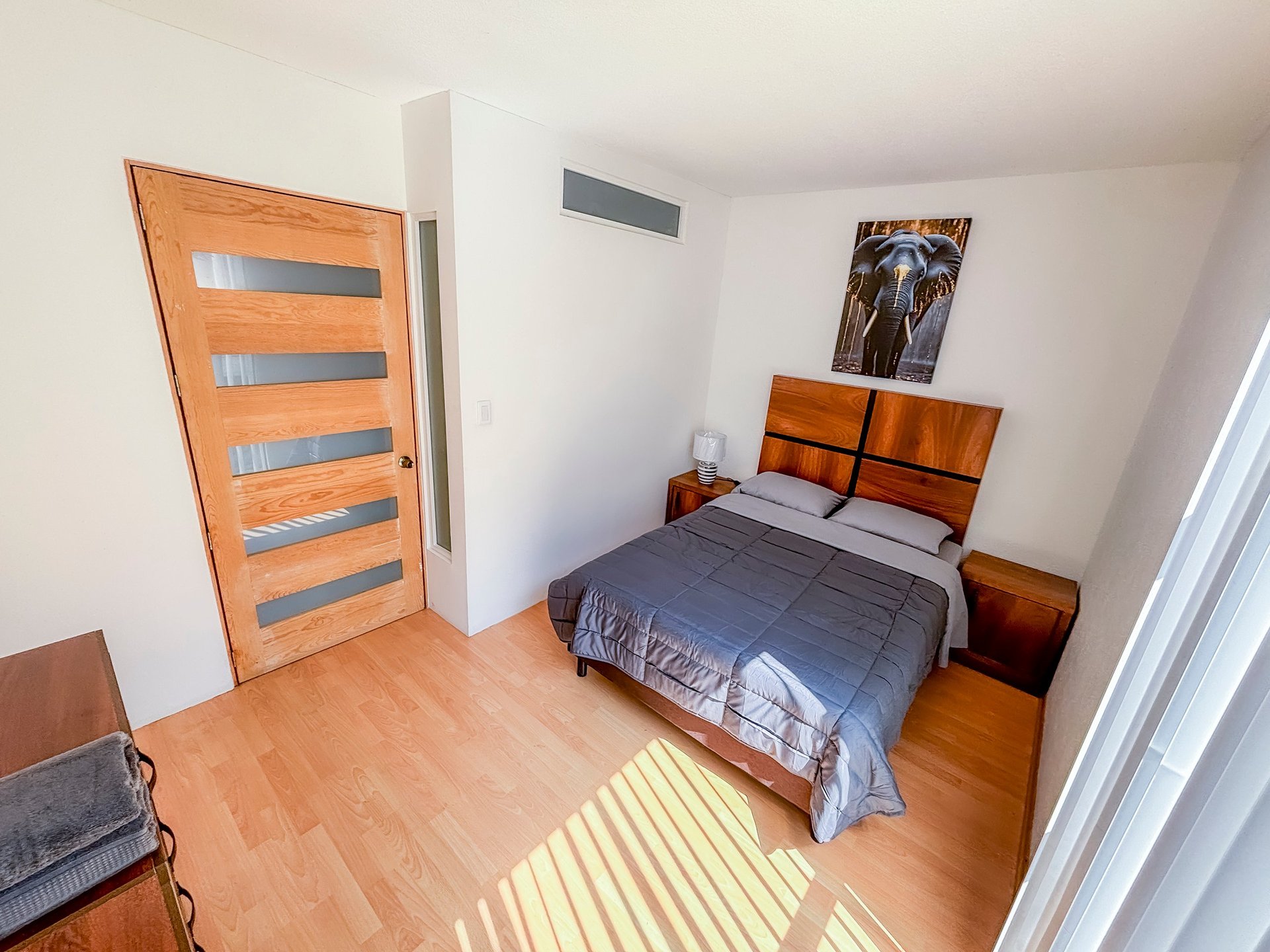 Minimalist bedroom with wooden flooring, orange door, blue-grey bed with wooden headboard, nightstands, and sunlight streaming through window