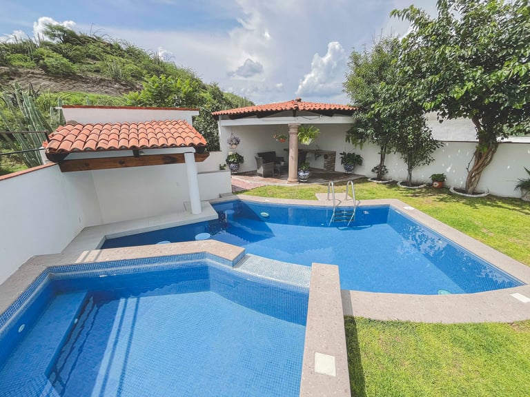 Modern backyard with two blue swimming pools, white walls with terracotta tile roof, lush green lawn, and surrounding vegetation under blue sky.