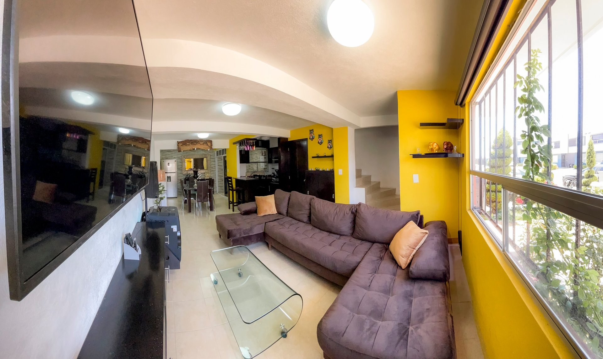 Modern living room with purple sectional sofa, yellow accent walls, bright ceiling lights, and large windows overlooking a city view