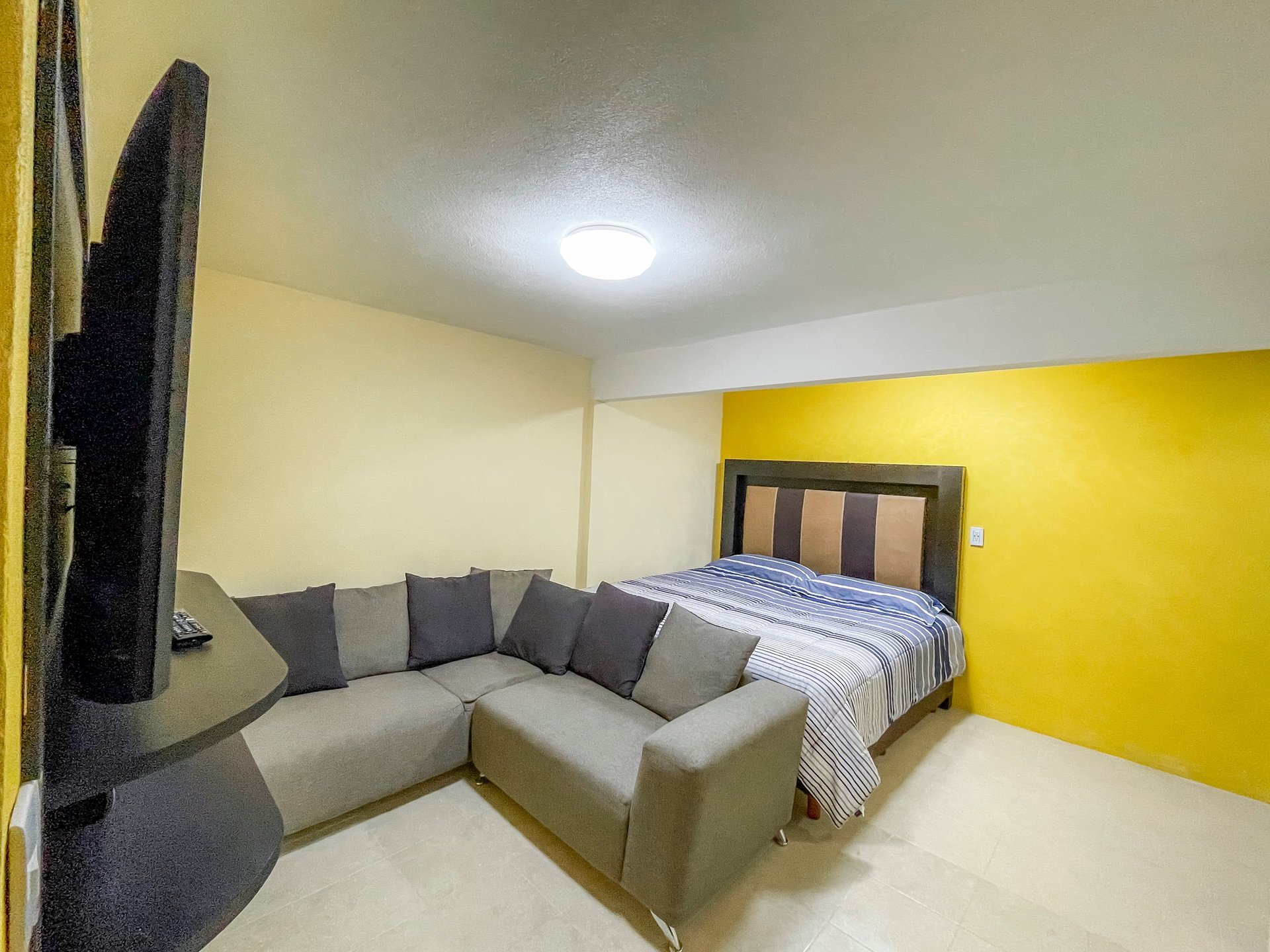 Modern bedroom with a gray sectional couch, bed with blue striped bedding, flat-screen TV on wall, and bright yellow accent wall