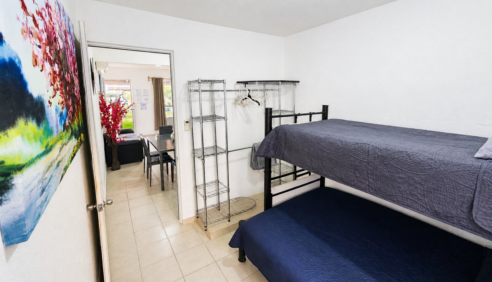 Bedroom with two beds, one black metal frame bed in foreground and one elevated bed in background, tile flooring, and abstract artwork on white wall.