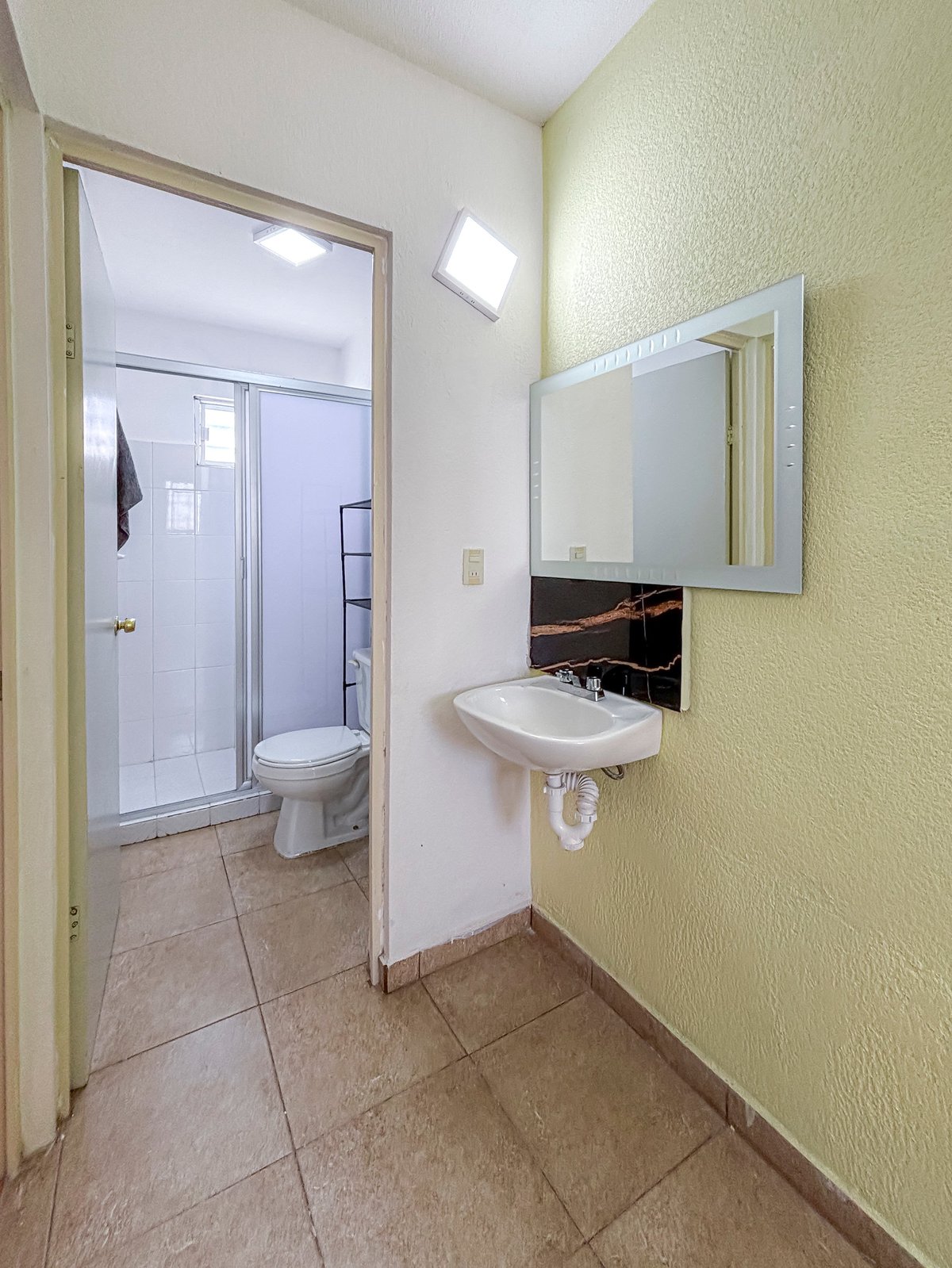 Modern bathroom with pale yellow walls, wall-mounted sink with mirror, and open doorway leading to shower stall with glass enclosure