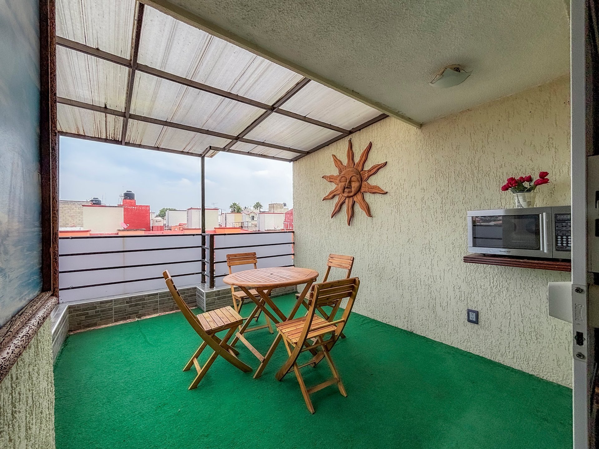 Modern rooftop terrace with green floor, wooden chairs, dining table, and city skyline view through glass railings.