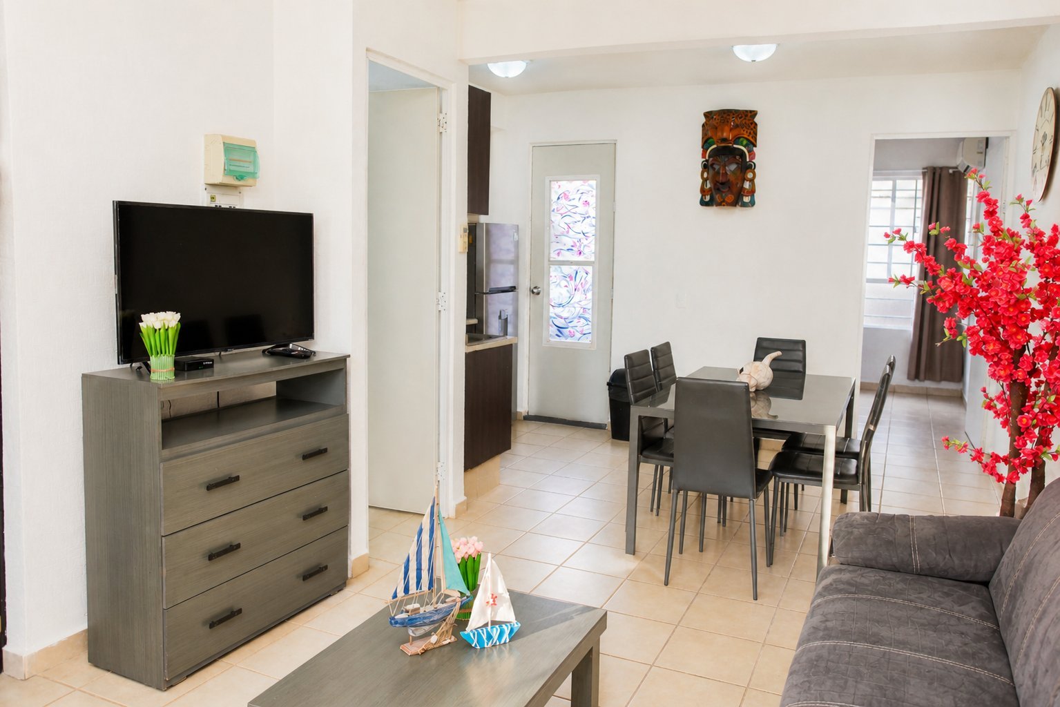 Modern open-plan apartment interior with living room featuring gray sofa, wooden TV stand, and dining area with black chairs and red flower arrangement.