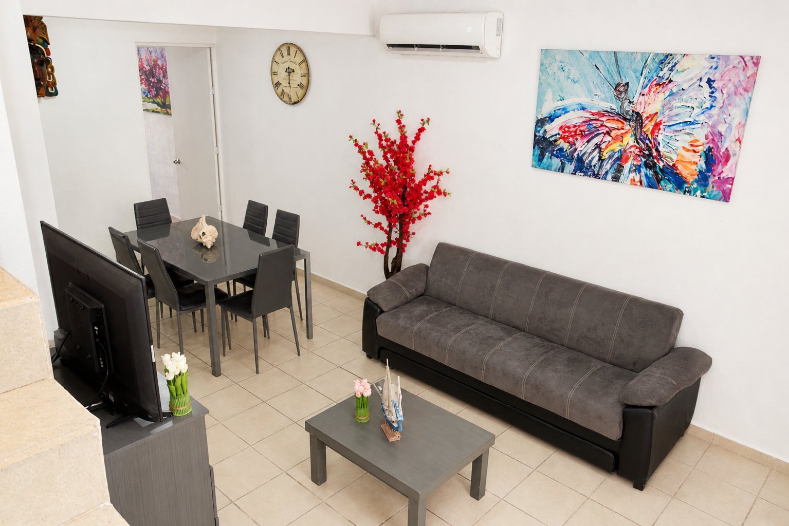 Modern living room with gray sofa, black dining table with chairs, coffee table, decorative artwork, red floral arrangement, and beige tile flooring.