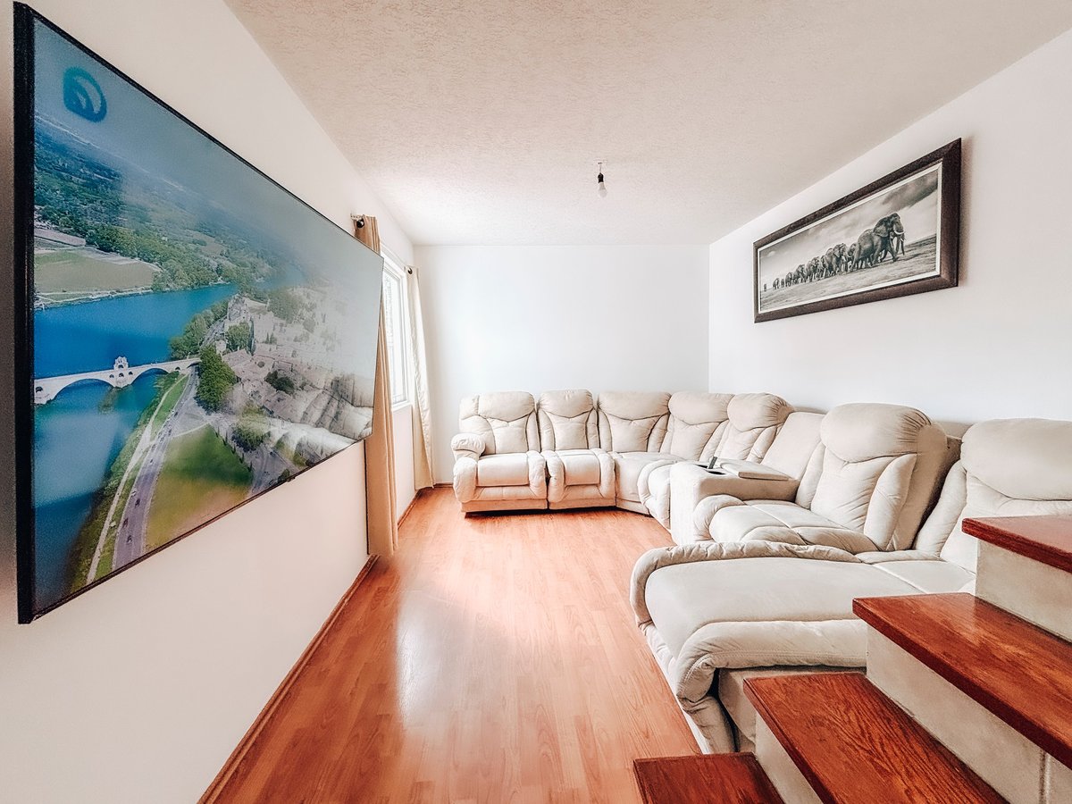 Modern living room with beige sectional sofa, wooden flooring, mounted TV displaying landscape, and framed wall art