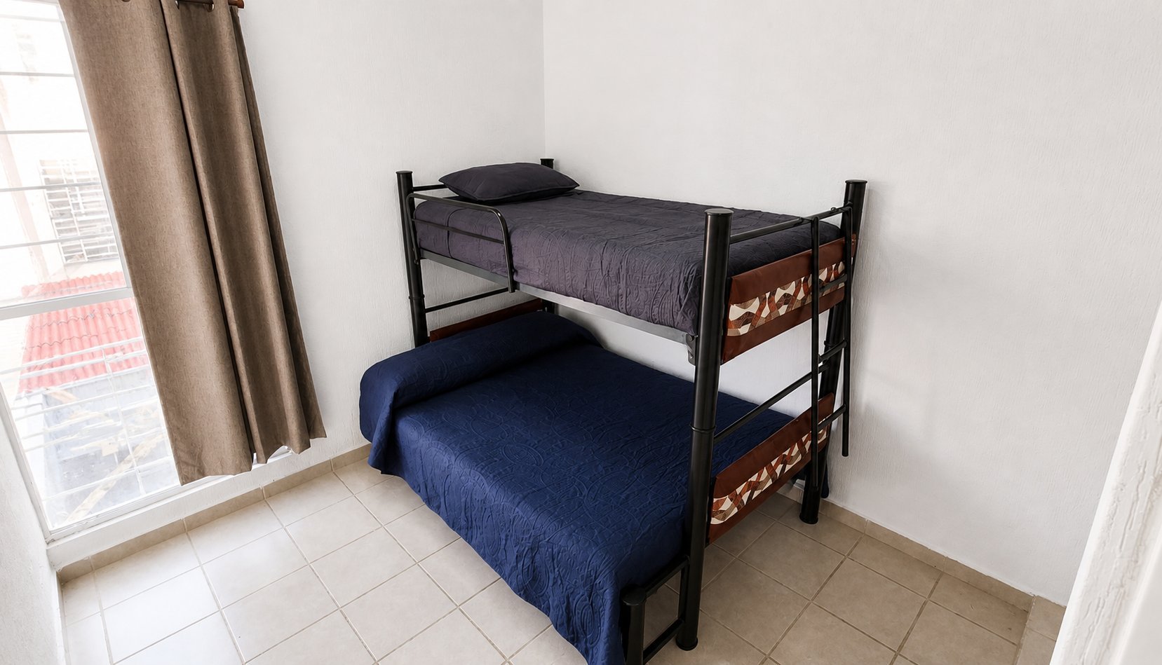 Black metal bunk bed with blue mattresses in a dorm room with tiled floor and window.