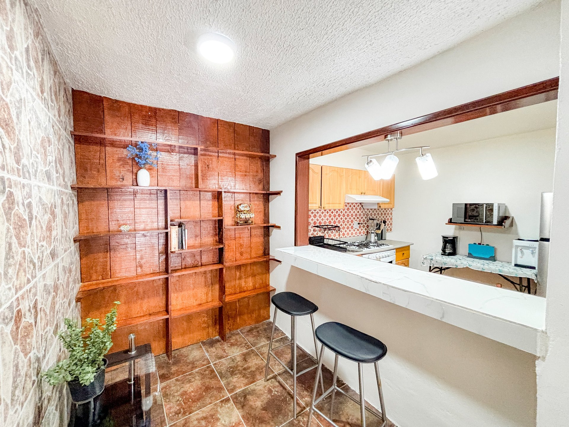 Modern kitchen with brown tile accents, bar seating with two black stools, and open pass-through window to adjacent room