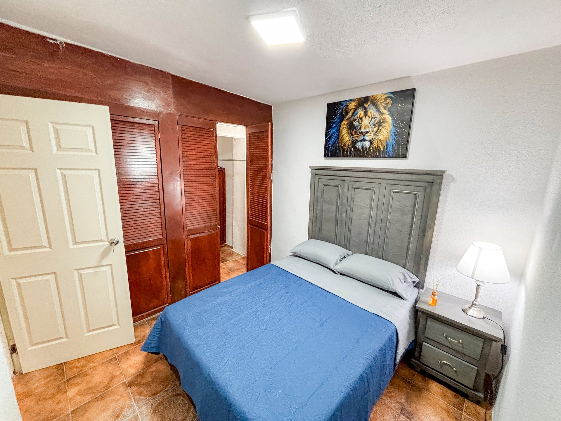 Bedroom with blue bed, gray headboard, nightstand with lamp, lion portrait above, red wood closet doors, and wooden floor