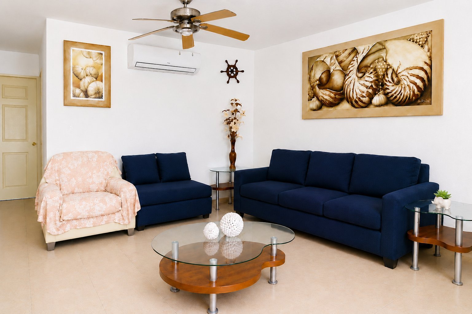 Modern living room with navy blue sofa and armchair, glass coffee table, framed artwork, ceiling fan, and nautical decor