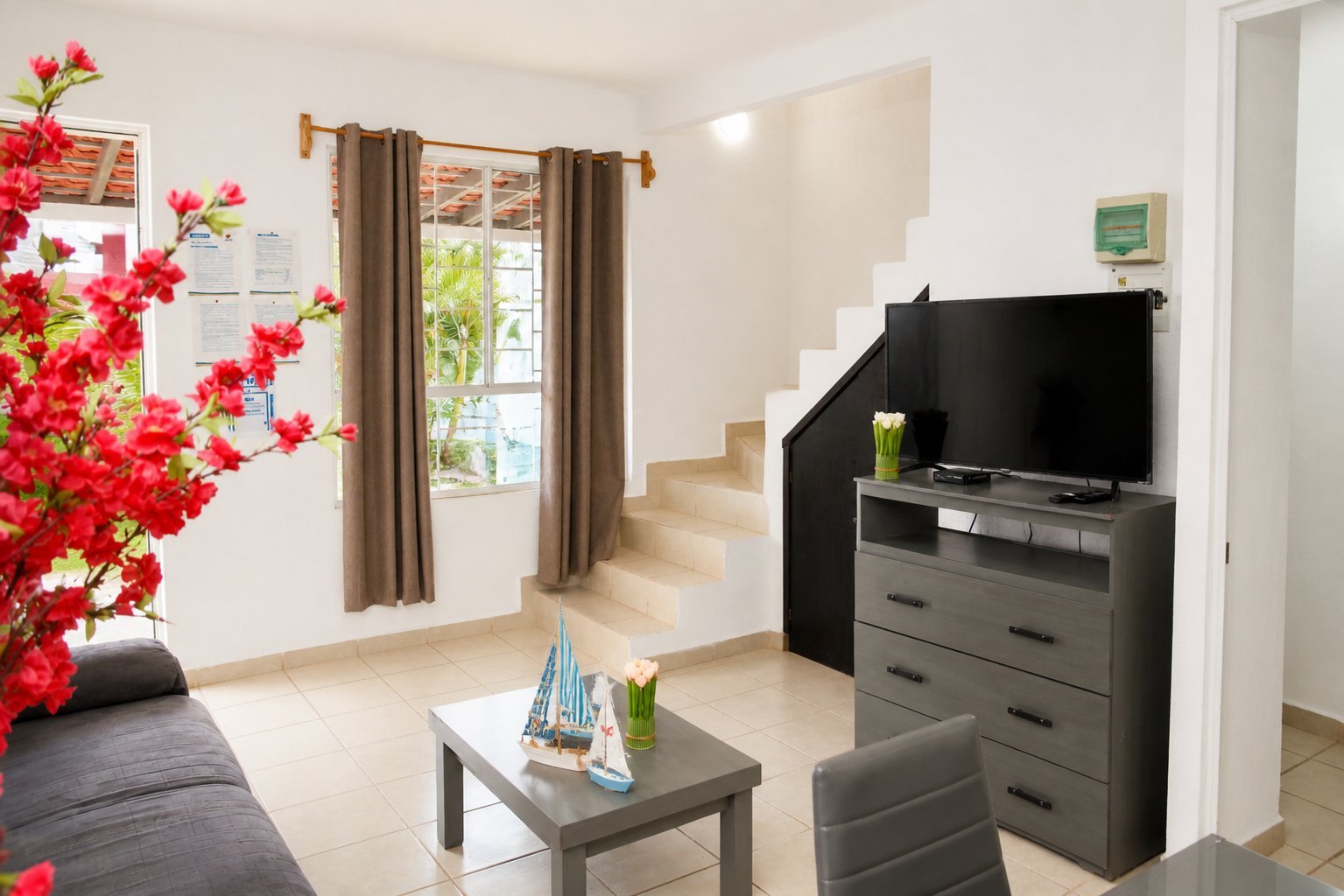 Modern living room with bright red flowers, flat-screen TV on black dresser, beige sofa, coffee table, and large windows with brown curtains.