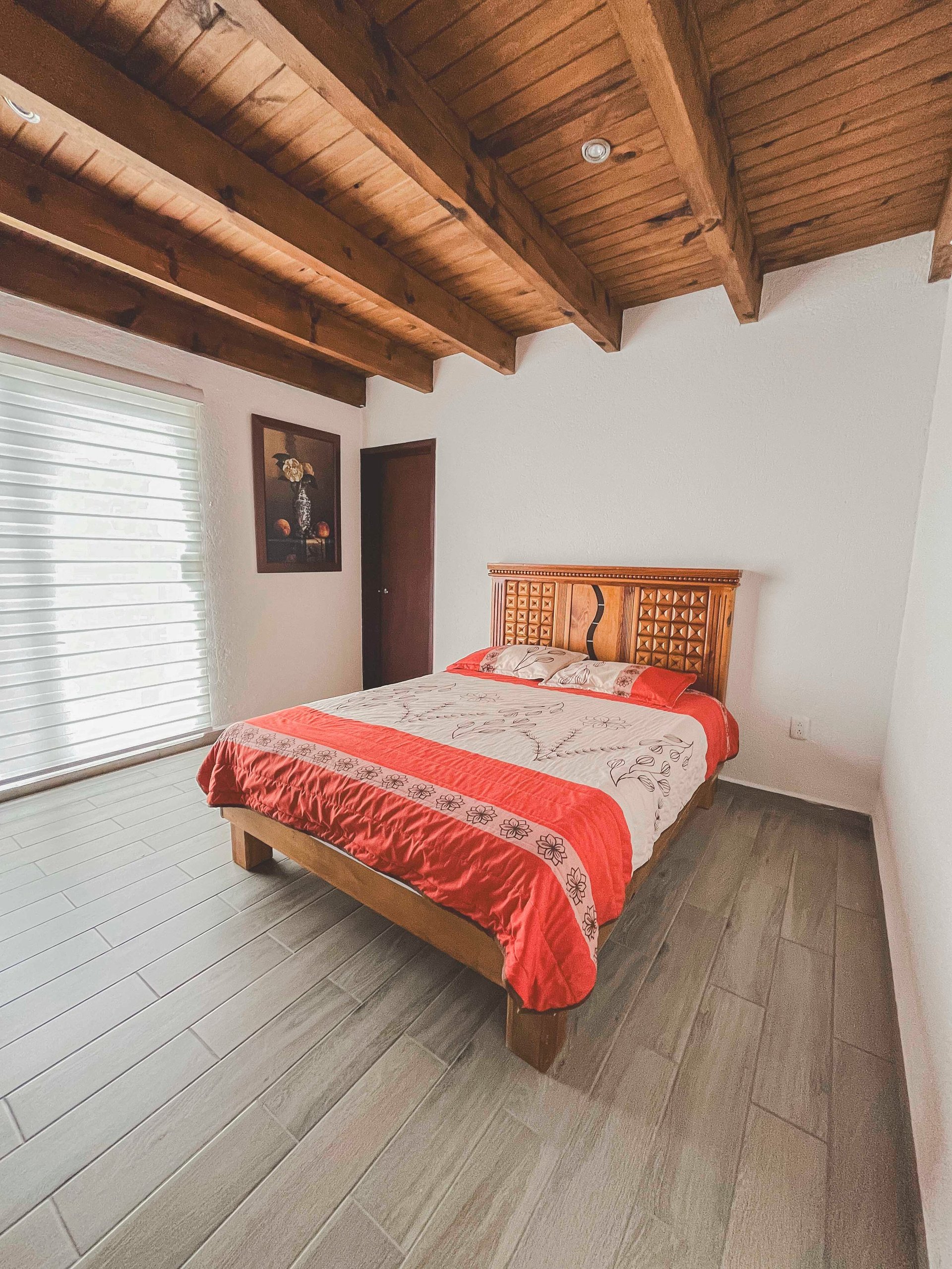 Modern bedroom with red and white bedding on wooden bed, exposed wooden ceiling beams, white walls, and wooden flooring